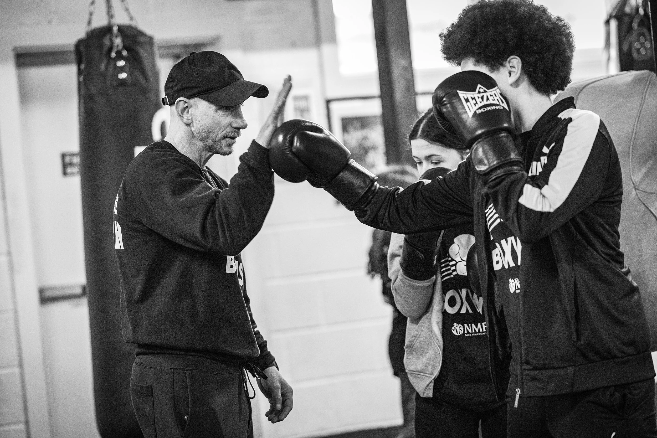 Boxing coach giving a high five to a female boxer in training, with another woman observing in the background, inside a gym.