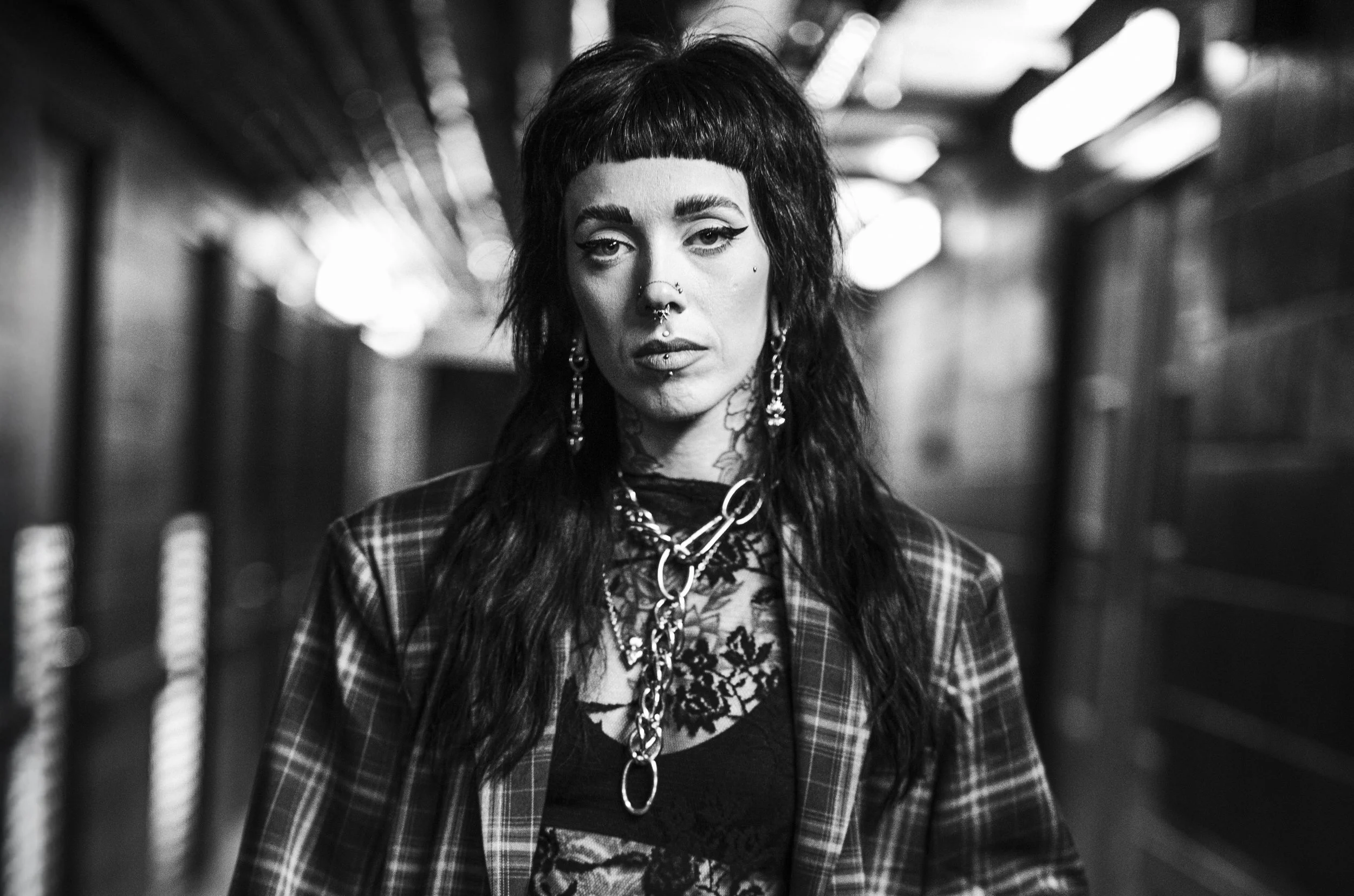 A black and white photograph of a woman with dark, shaggy hair and multiple facial piercings, wearing statement jewelry and a checkered jacket, standing in a dimly lit, industrial-style hallway.