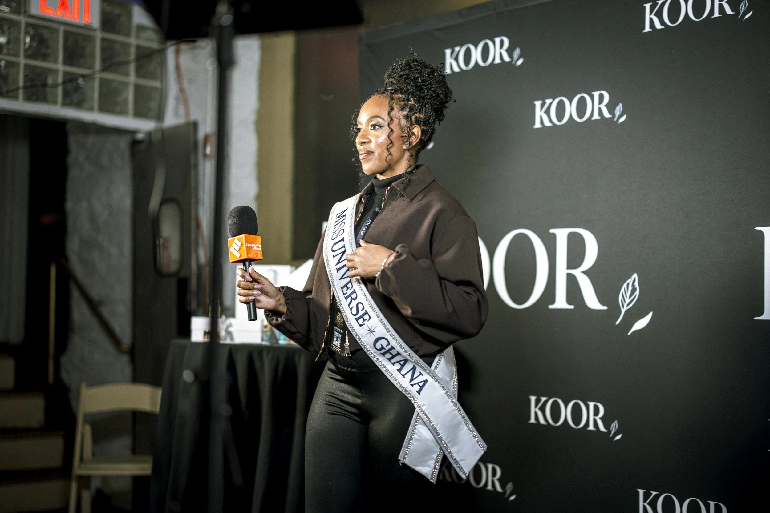 A woman wearing a sash that reads 'Miss Universe Ghana' holding a microphone in front of a black background with white text and leaves motif, at an event or interview setup.