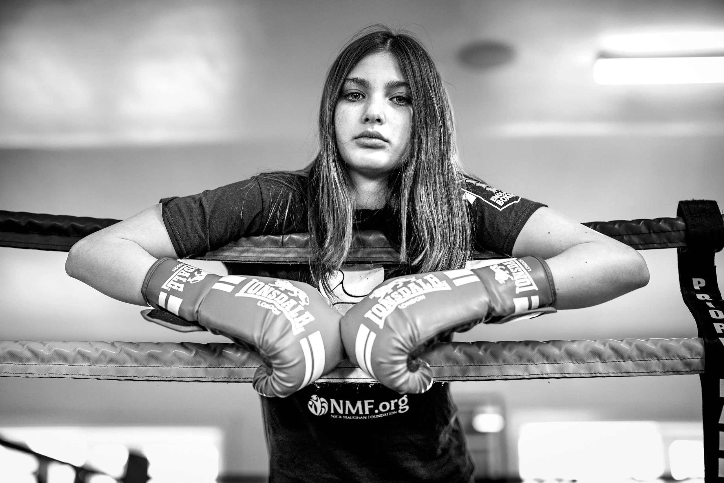 A female boxer leaning on the ropes of a boxing ring, wearing boxing gloves and sportswear, looking directly at the camera with a serious expression.