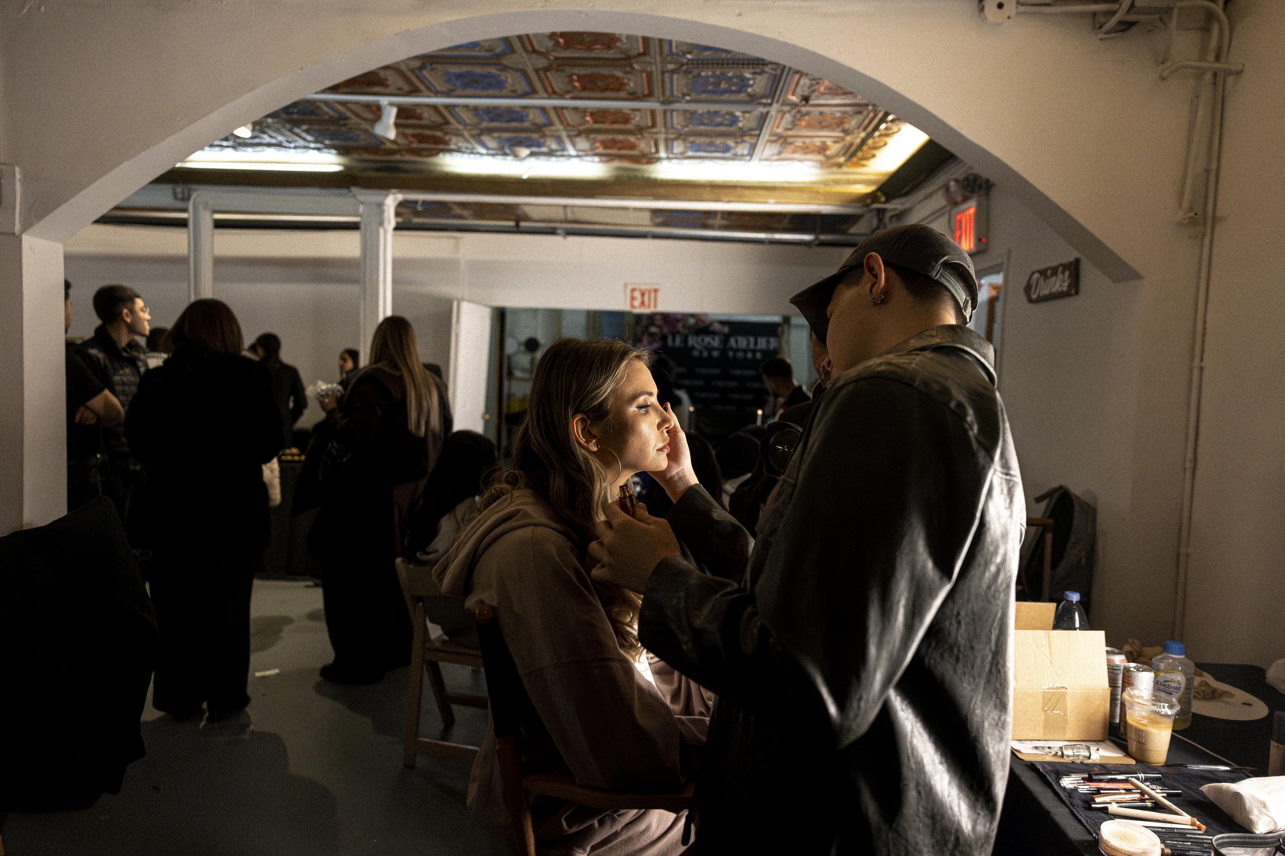 A woman gets her makeup applied by a makeup artist in a busy indoor setting, with other people and tables visible in the background.