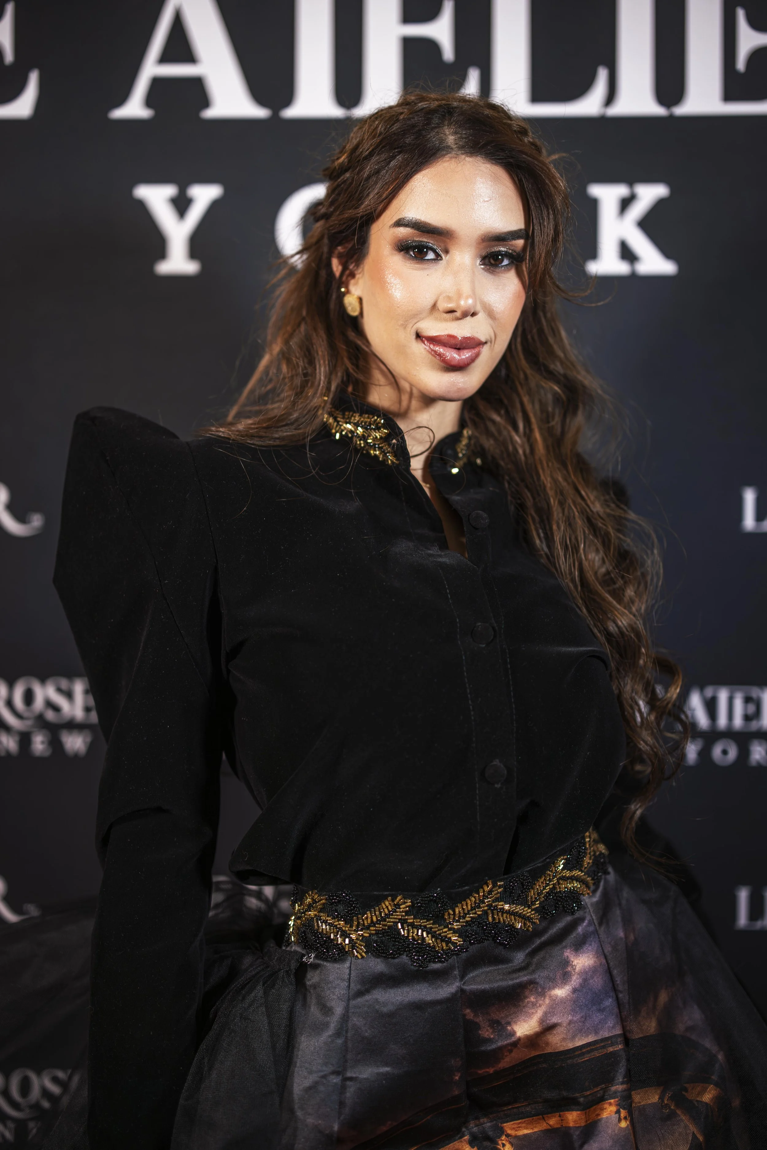 A woman with long wavy brown hair and makeup, wearing a black jacket with gold embellishments, standing in front of a dark background with white text.