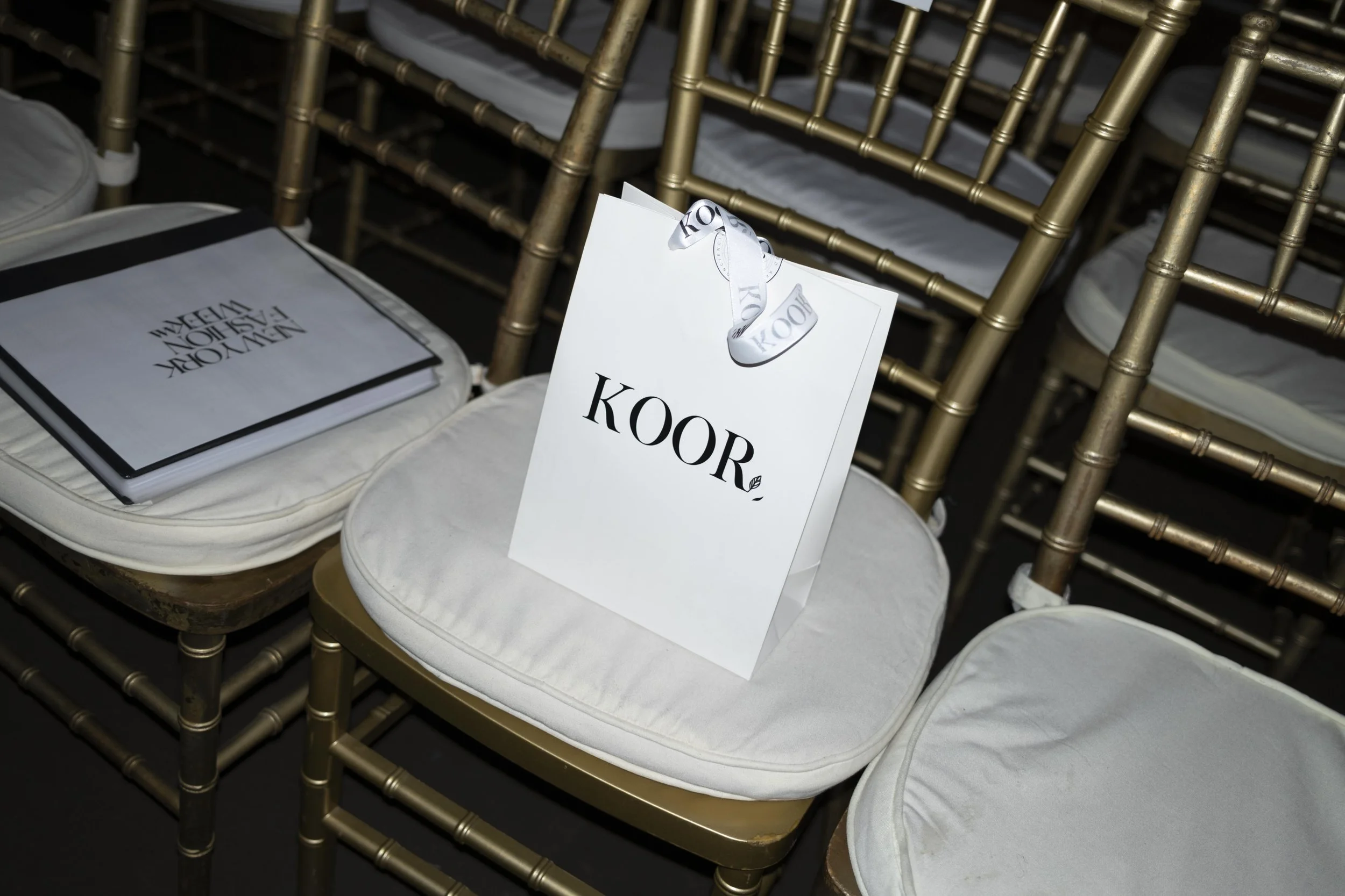 Multiple gold-colored chairs arranged in rows, with white cushions on the seats. A white gift bag with black lettering and a ribbon is placed on one of the chairs, and a white folder or program with black text is also on a chair.