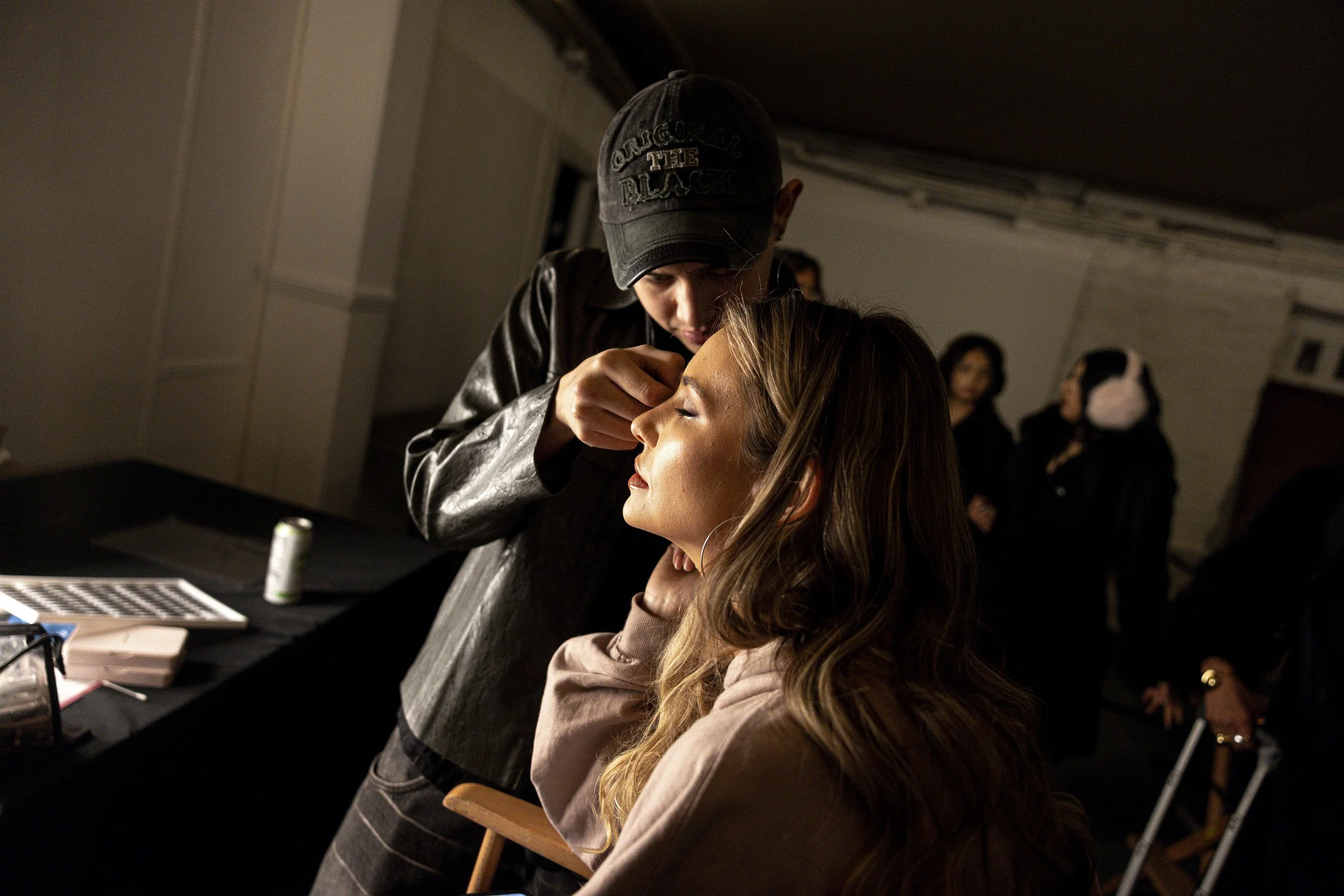 A makeup artist is applying makeup to a woman with long, wavy hair who is sitting calmly with her eyes closed. The artist is wearing a black gymnast cap and a leather jacket, focusing on her work. In the background, three women are talking and watchi