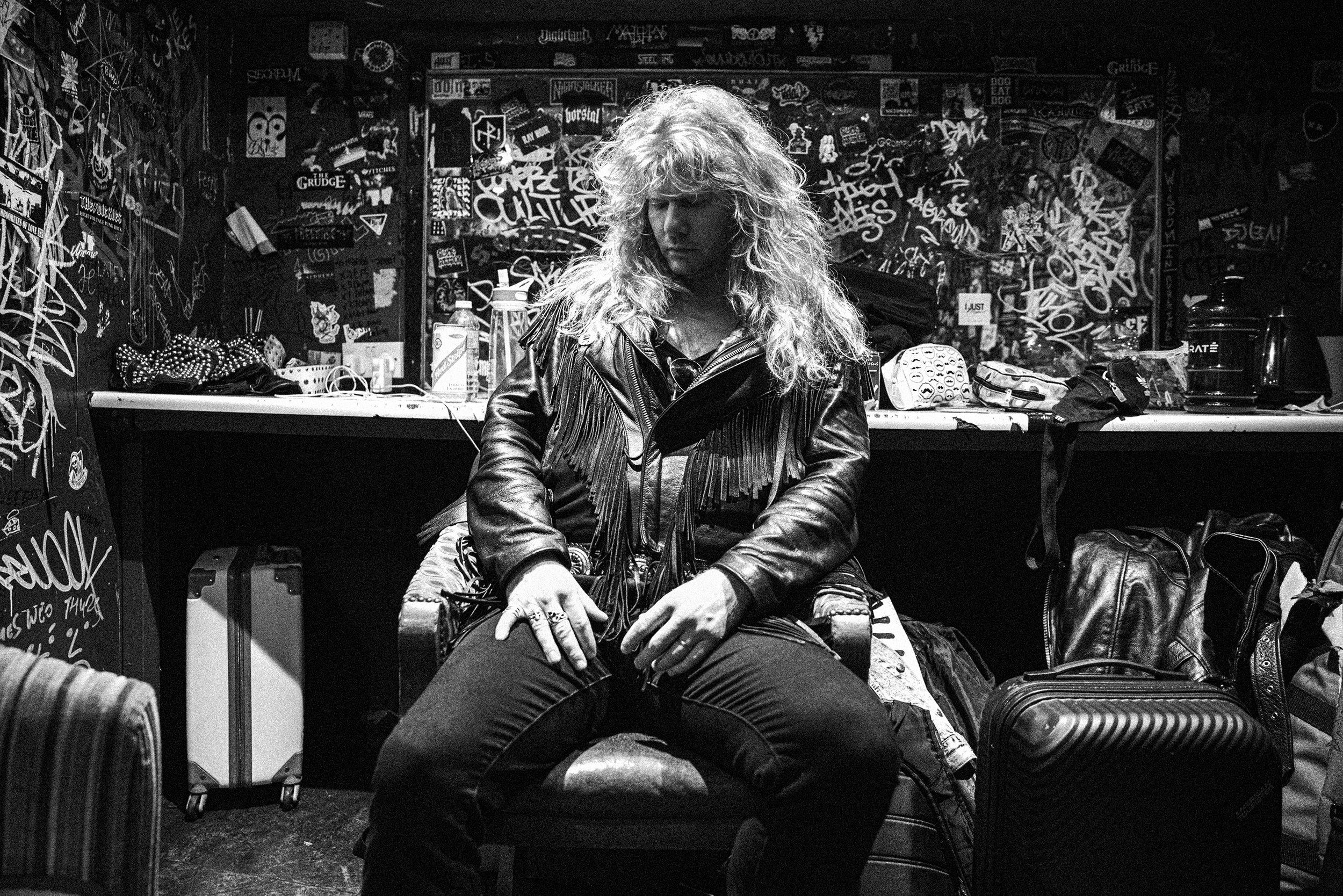 A woman with long curly hair sitting on a chair in a music or art studio filled with graffiti and stickers on the walls, dressed in a leather jacket and jeans.