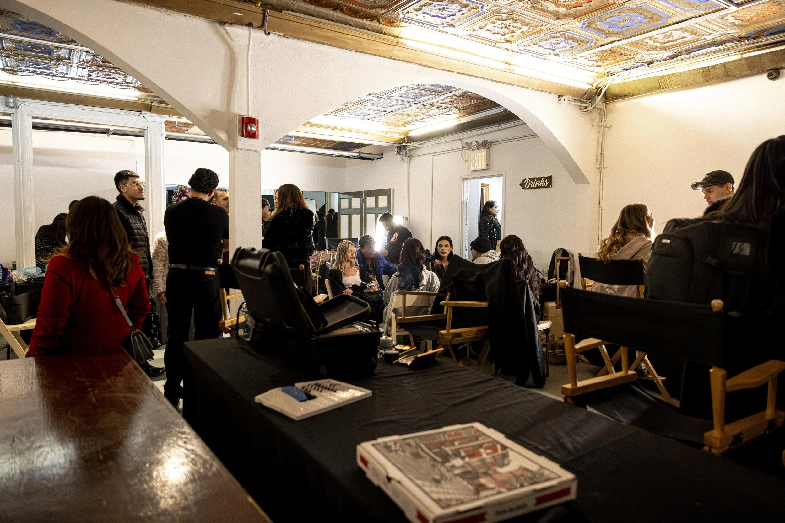Group of people gathered in a room with a colorful, patterned ceiling and white walls, some standing and some sitting, with a table and chairs nearby. There is a sign that says 'Drinks' on the wall and an exit door.