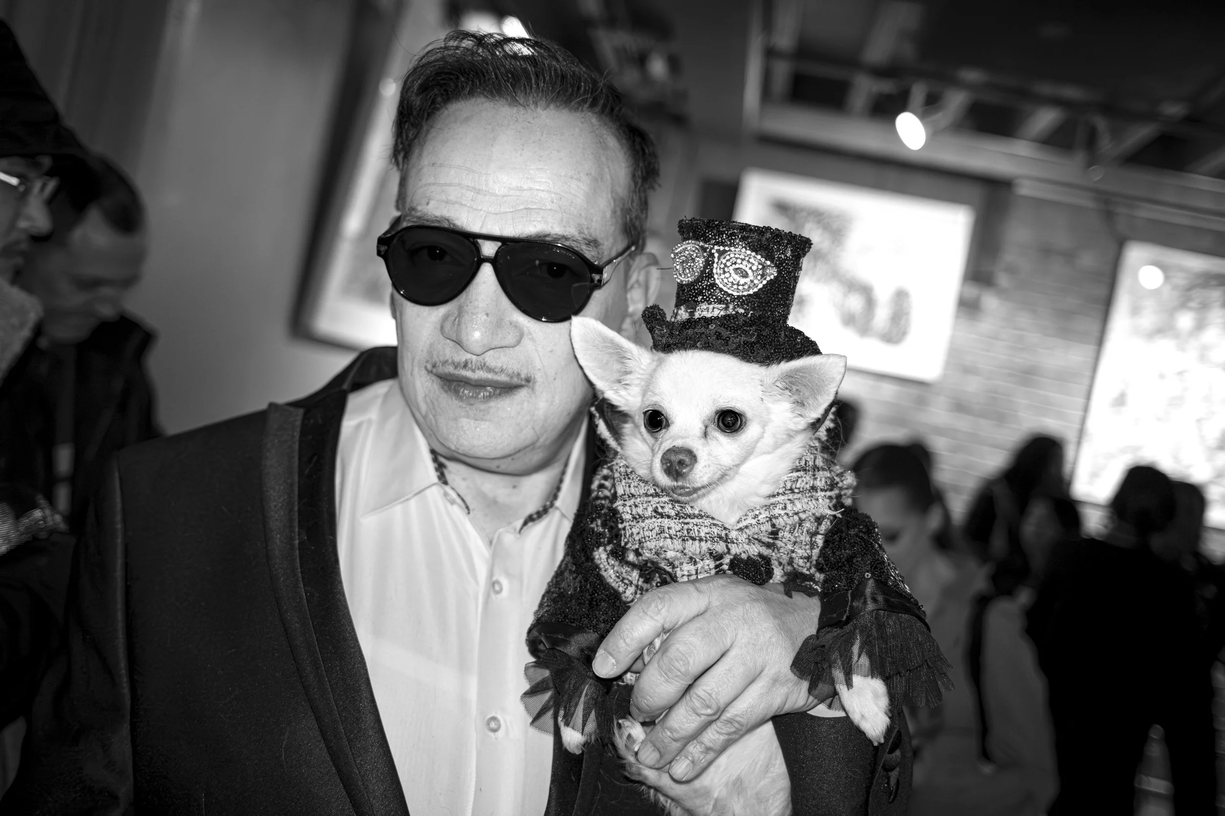 A man with slicked-back hair wearing dark sunglasses and a white shirt, holding a small dog dressed in a costume and a tiny hat, in an indoor setting.