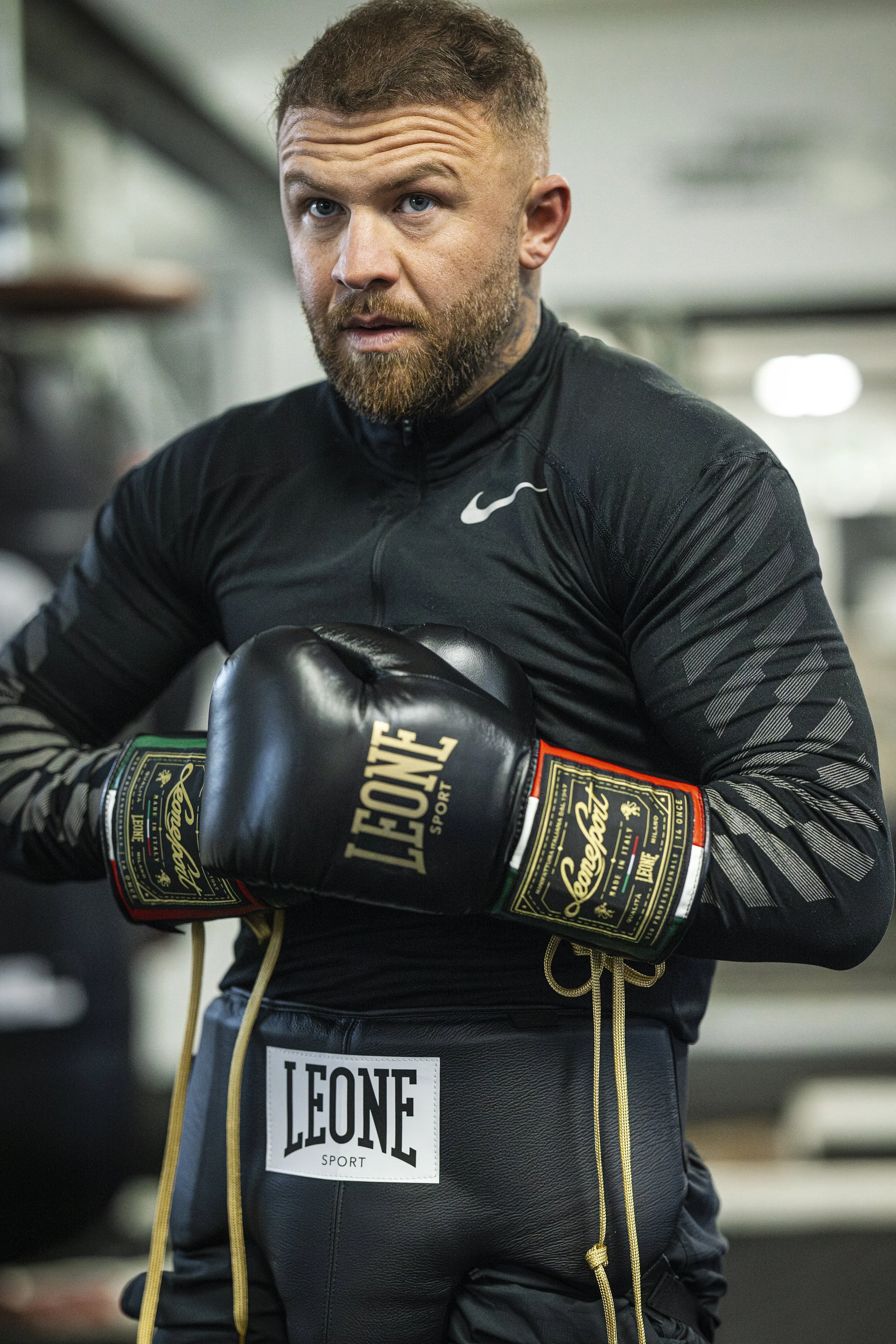 A man with a beard and short hair in a black athletic jacket is wearing boxing gloves and posing in a gym.