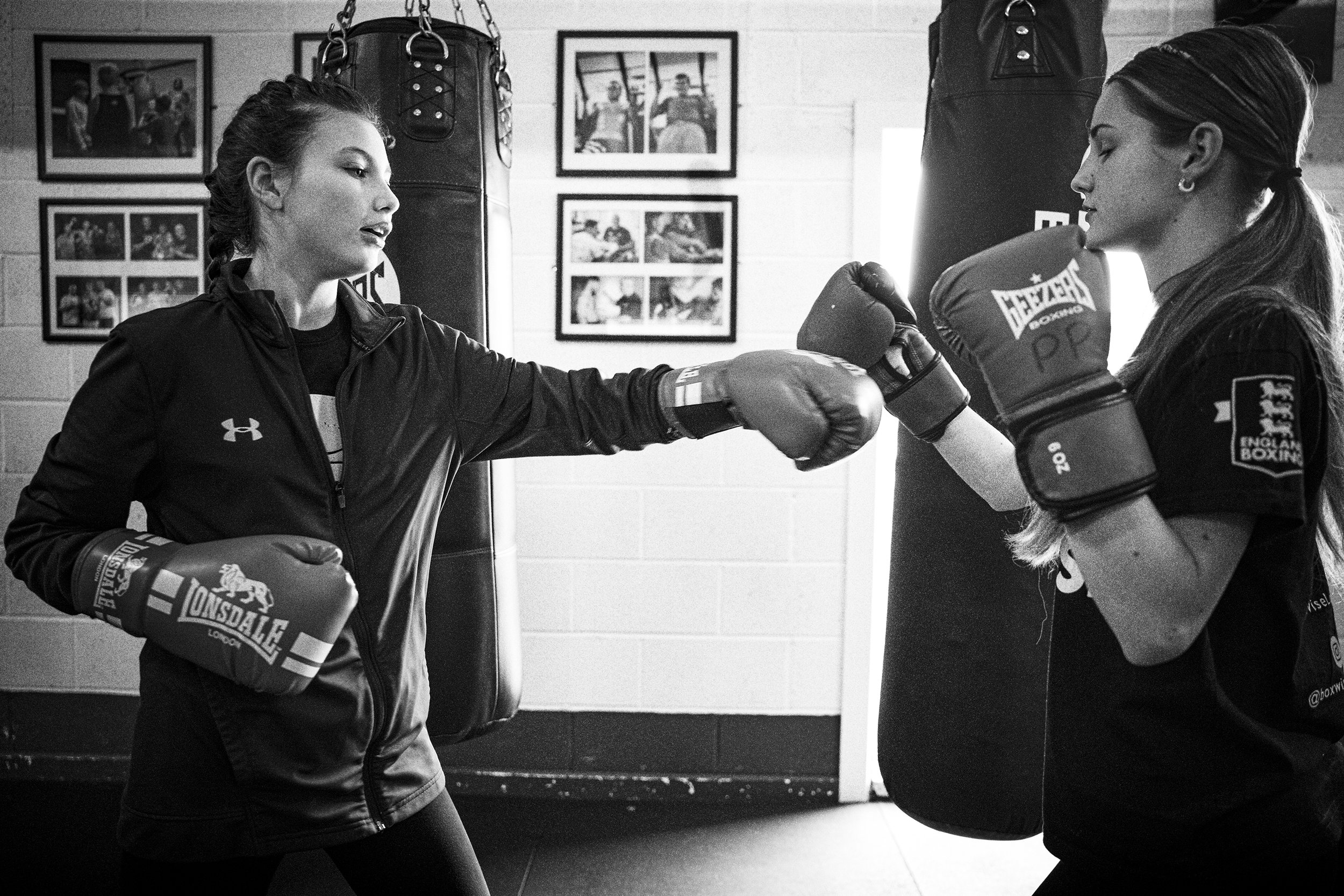 Two women practicing boxing in a gym, one throwing a punch while the other blocks with her gloves, with punching bags and framed photos on the wall in the background.