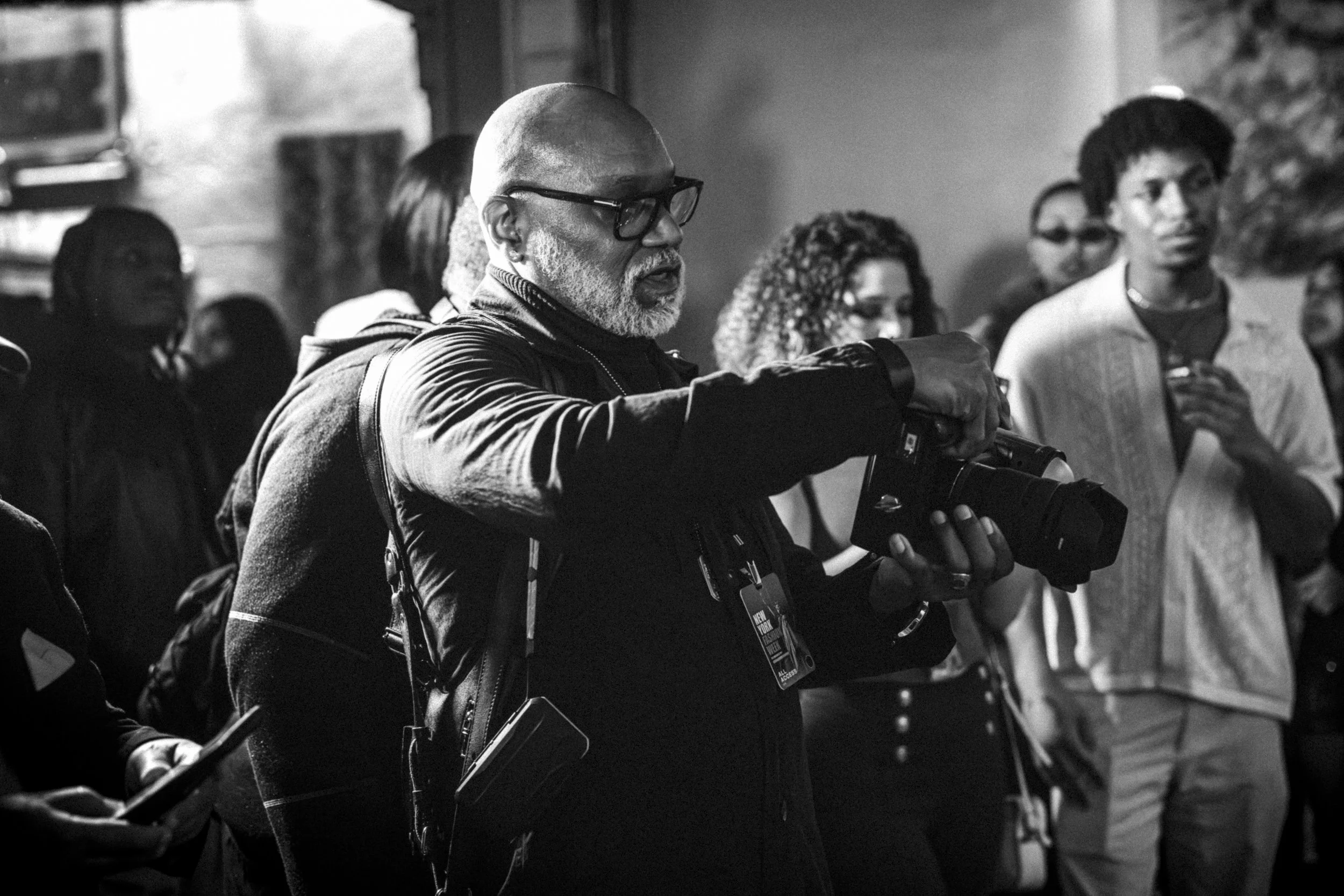A diverse group of people standing together in an indoor setting, with one man in the foreground holding a camera. The scene is in black and white.