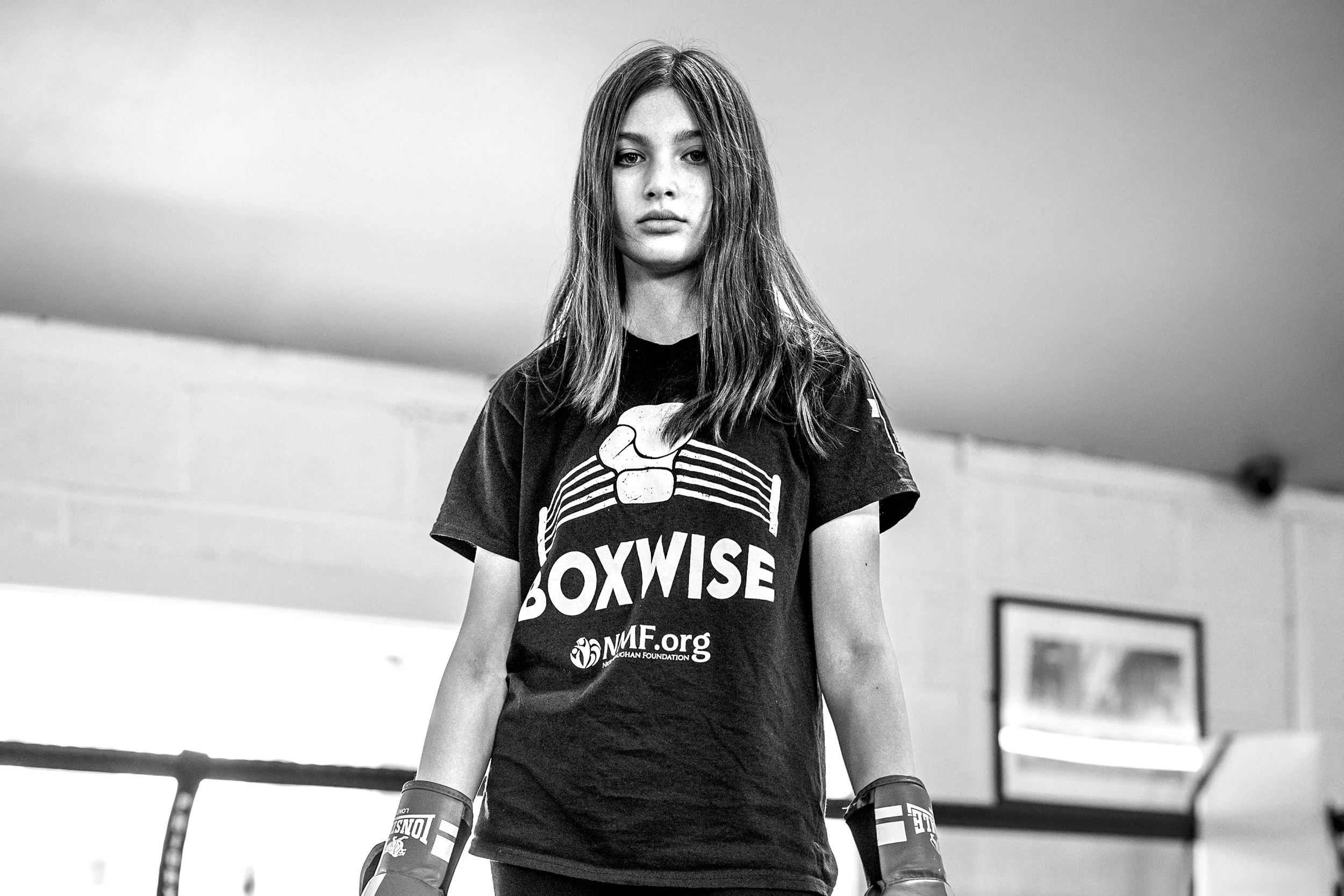 Black and white photo of a young female boxer at a gym, wearing boxing gloves and a t-shirt with the text 'Boxwise' and a logo, standing in a boxing ring.