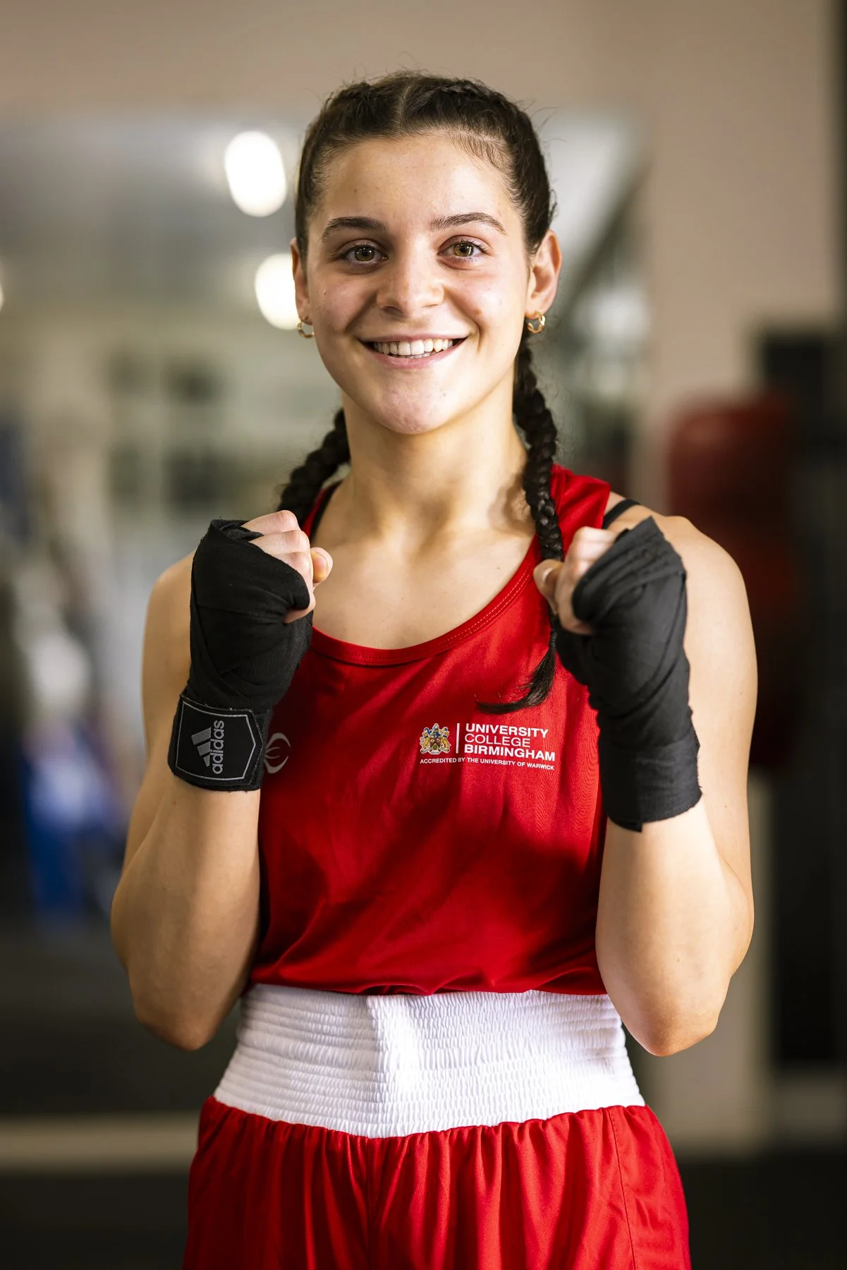 A young female boxer wearing a red tank top and shorts, with boxing gloves, smiling confidently in a gym.