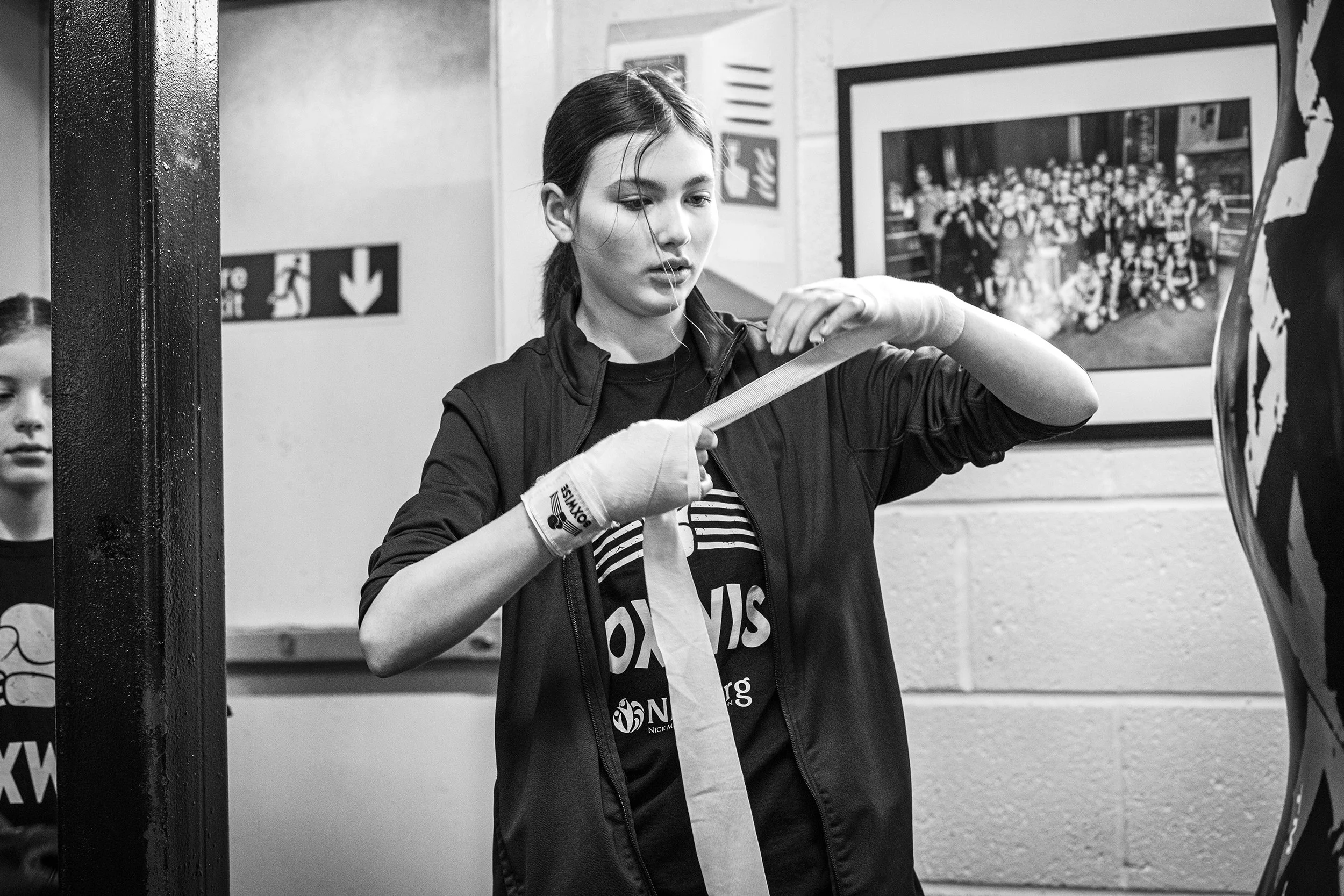 A young girl in a black jacket wrapping her hands with sports tape in a gym or training area, with a painting or photograph of a group of people in the background.