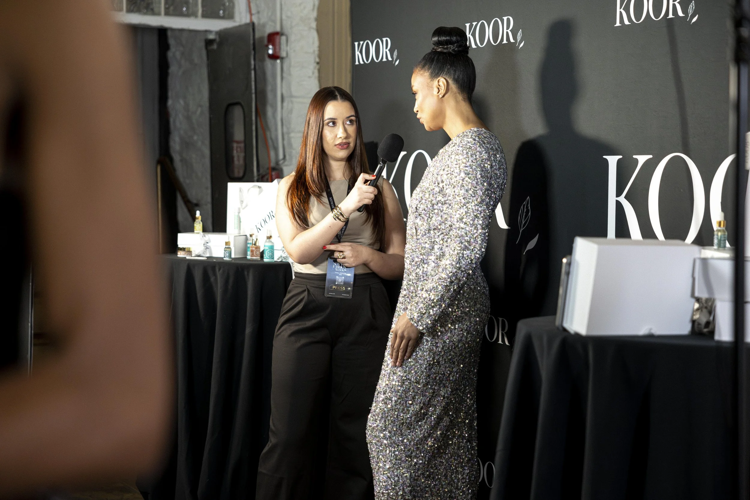A woman being interviewed with a microphone by another woman at an event. The background has the word 'KOOR' repeatedly written on it, and there is a table with various small bottles and products.