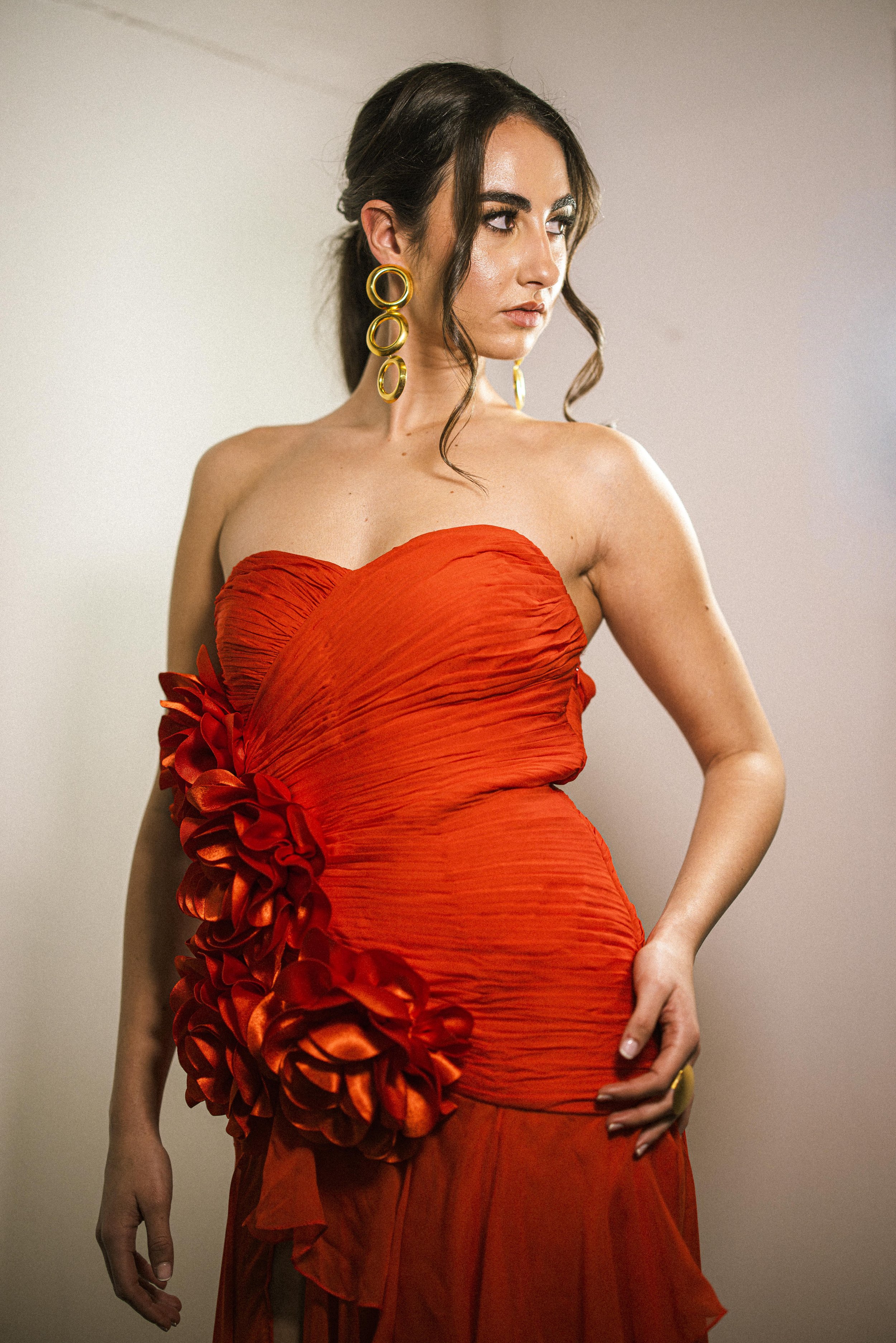 A woman wearing a strapless red dress with ruffles and large gold hoop earrings posing against a plain background.