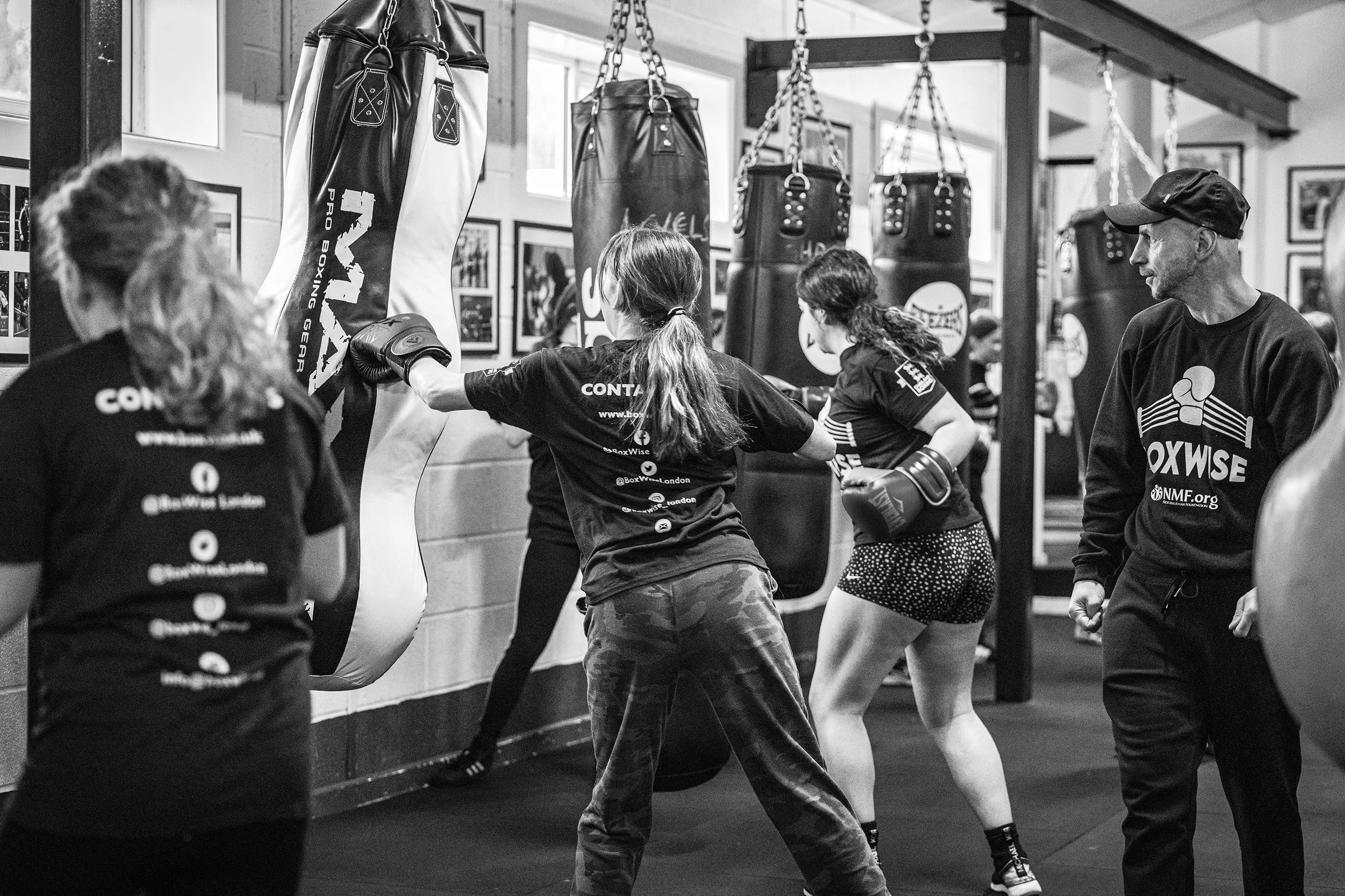 Boxing class with women practicing punches on heavy bags at a gym, supervised by a trainer.