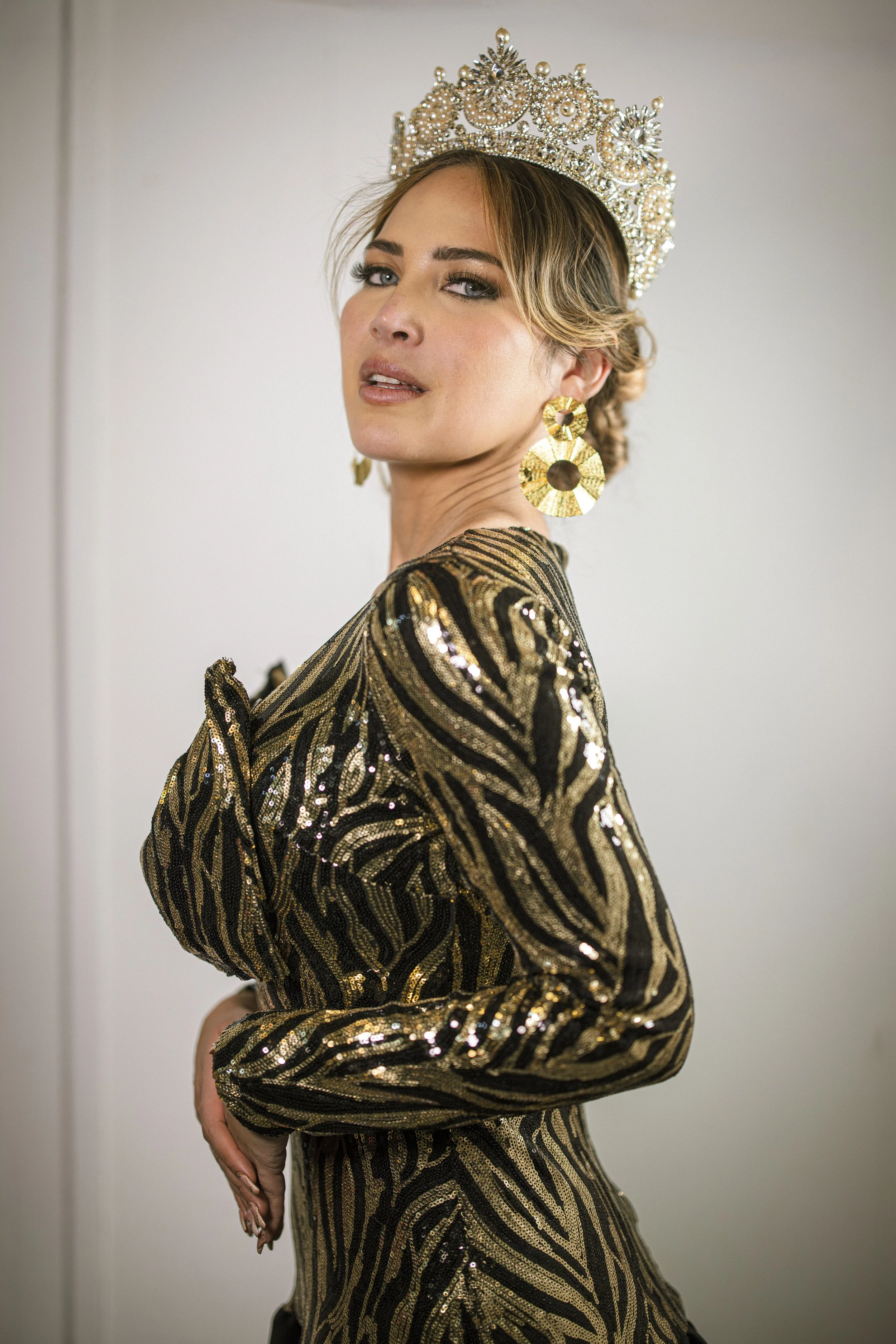A woman wearing a gold and black striped, sequined dress with large gold earrings, a crown, and makeup. She is posing with her right hand on her waist and her head turned slightly to the left.