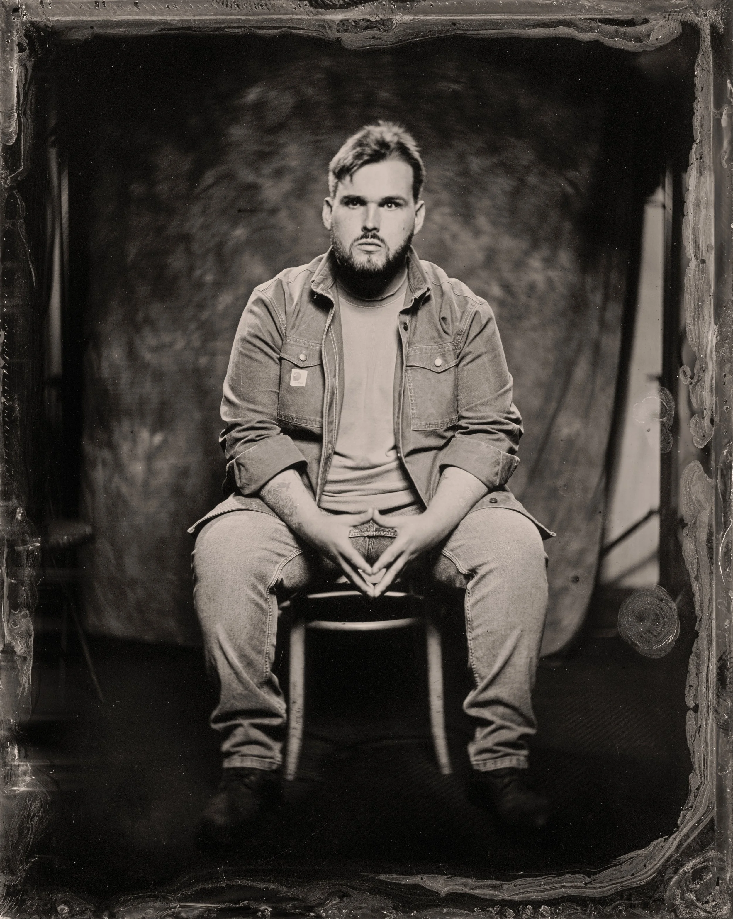 Black and white portrait of a young man with beard and short hair, wearing a denim jacket over a T-shirt, sitting with his hands clasped, in front of a dark, textured background.