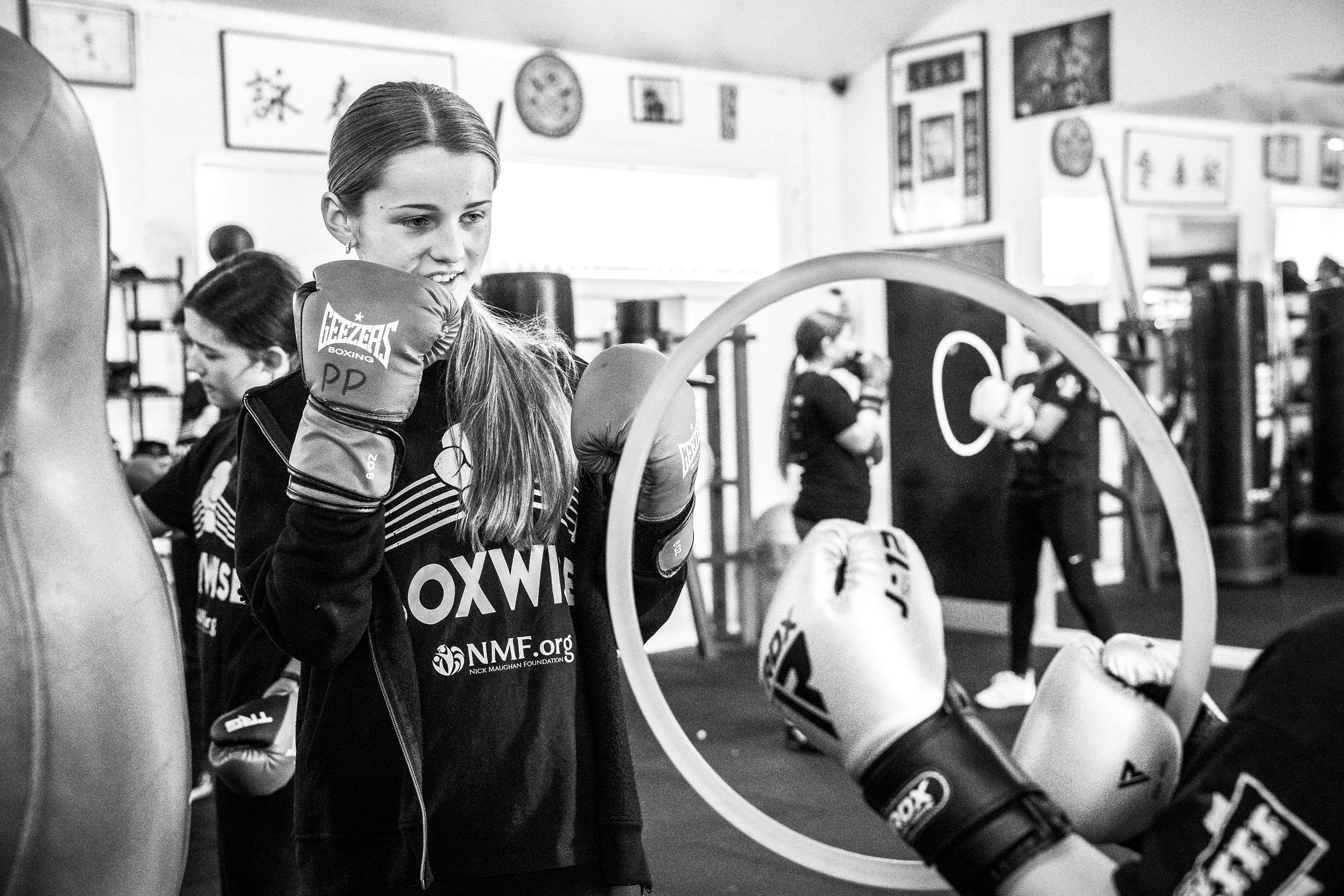 Young woman in boxing gloves practicing in a gym with other women boxing in the background.