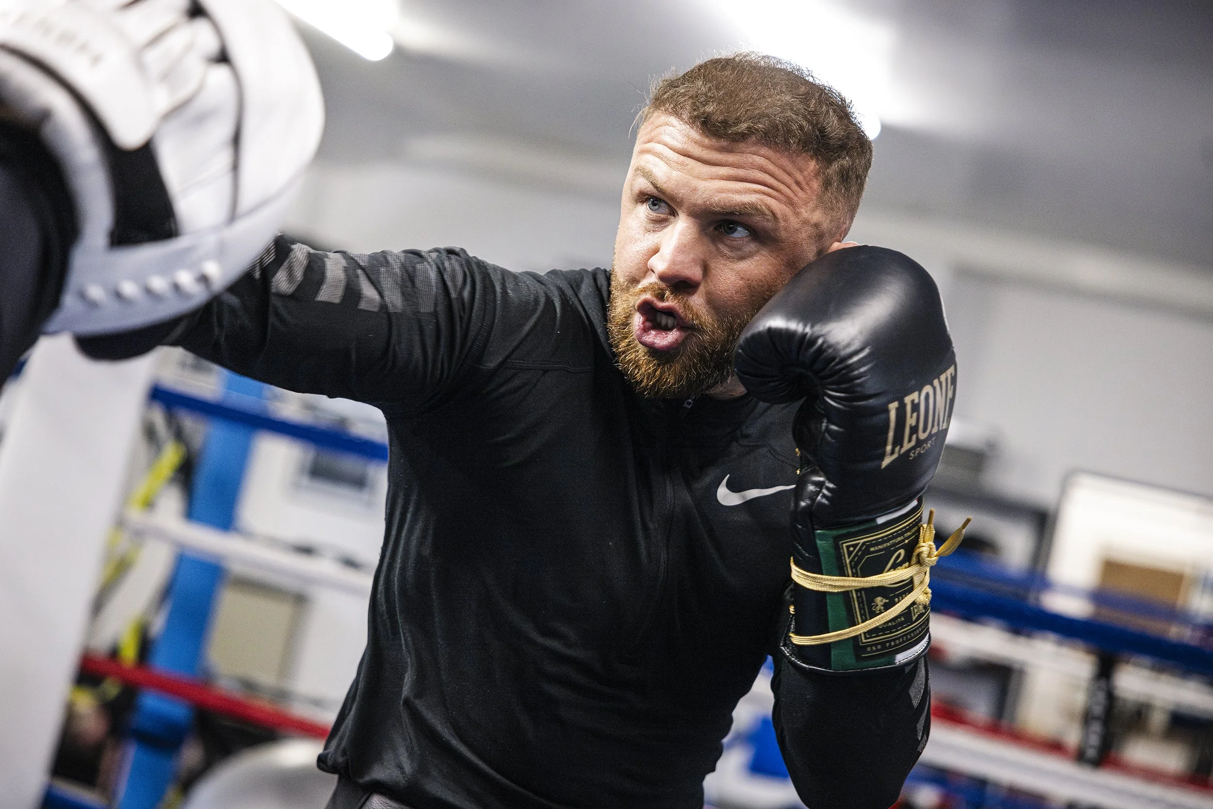 A male boxer with a beard, wearing black athletic gear and black boxing gloves, is throwing a punch at a boxing training mitt in a gym.