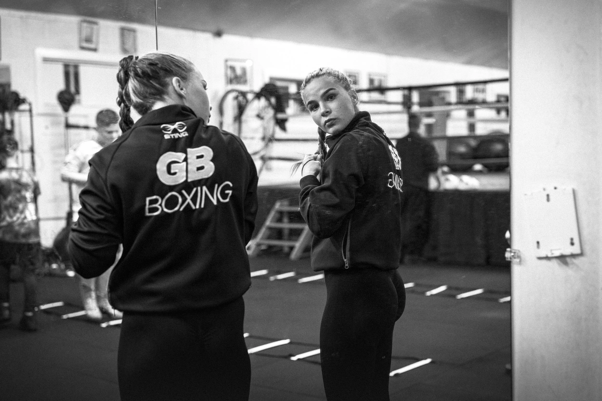 Two young women in boxing jackets at a gym, one looking at the camera, the other with her back turned.