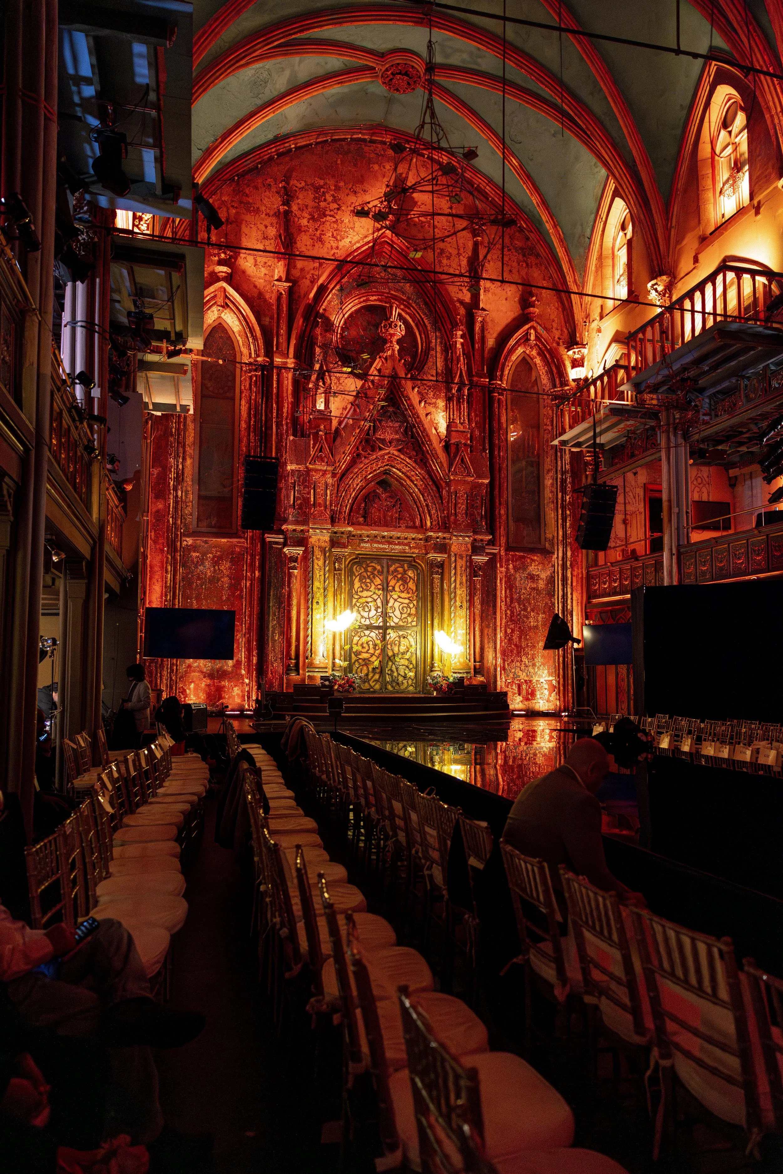 Interior of a Gothic-style church or cathedral with ornate architecture and high arched ceilings illuminated in warm orange lighting, with chairs lined up for an event or performance.