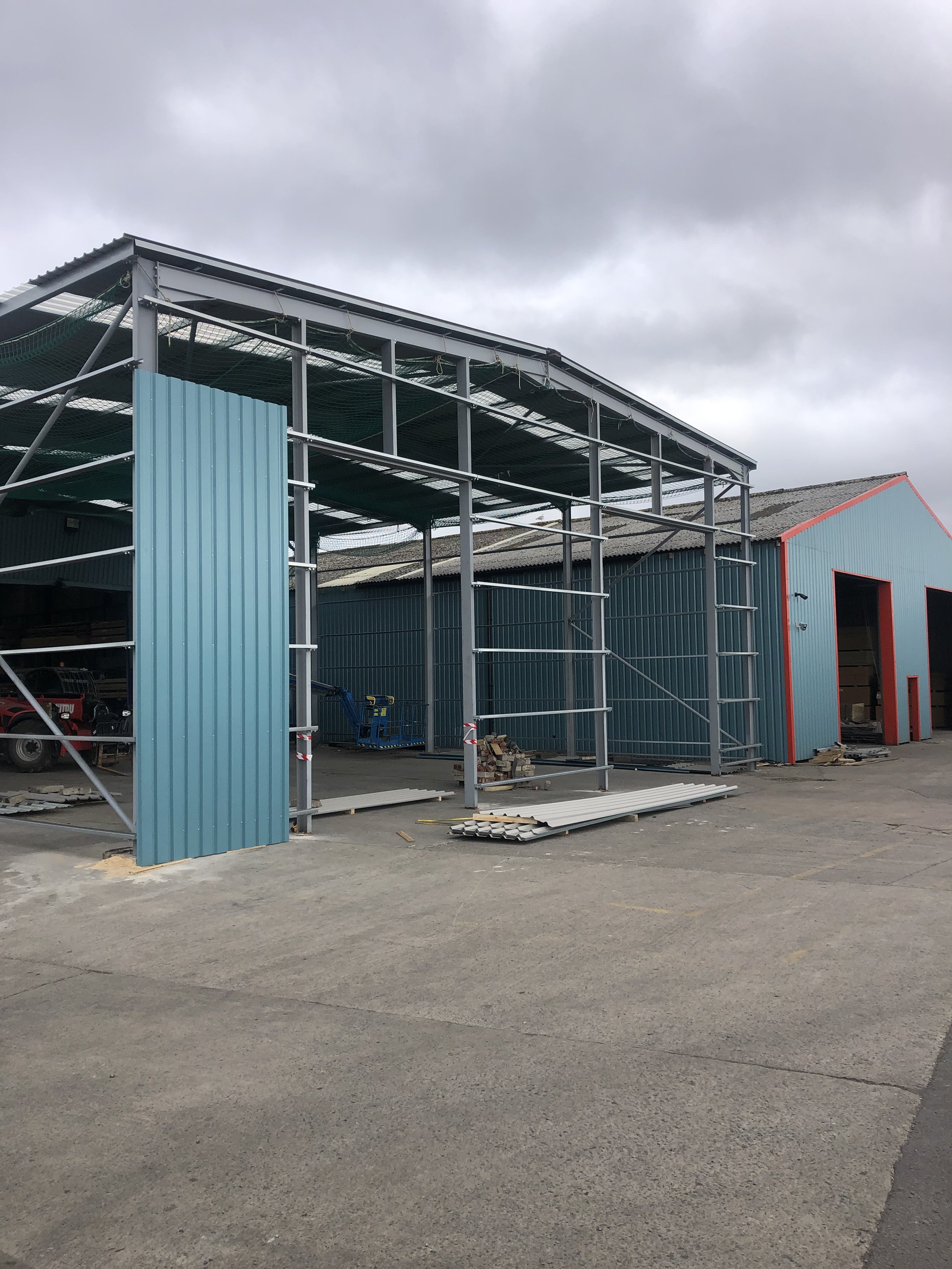 Partially constructed steel frame building next to a completed industrial warehouse, with construction materials and equipment on the ground.