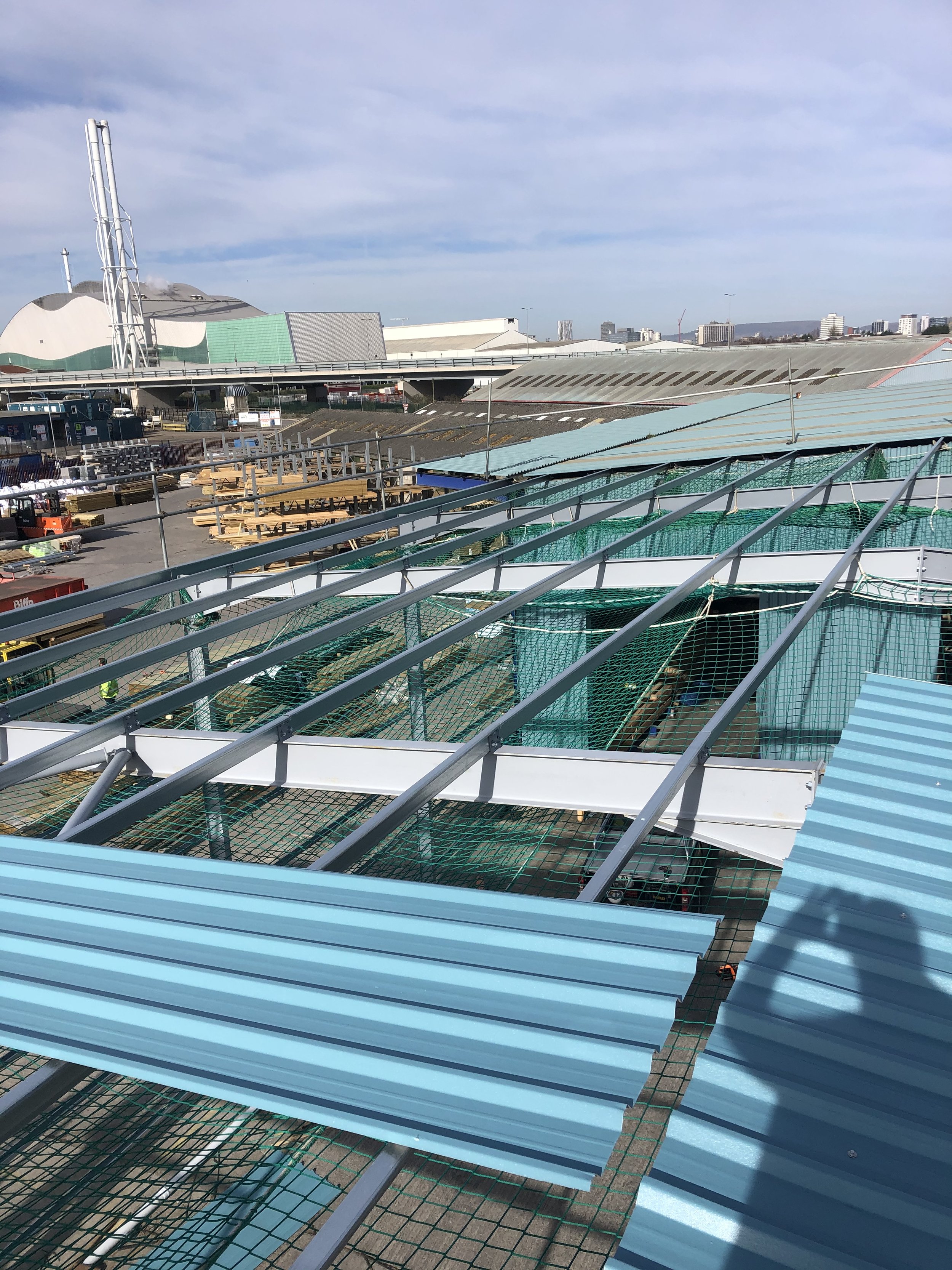 Construction site with metal framework and blue roof panels, industrial background with buildings and structures.