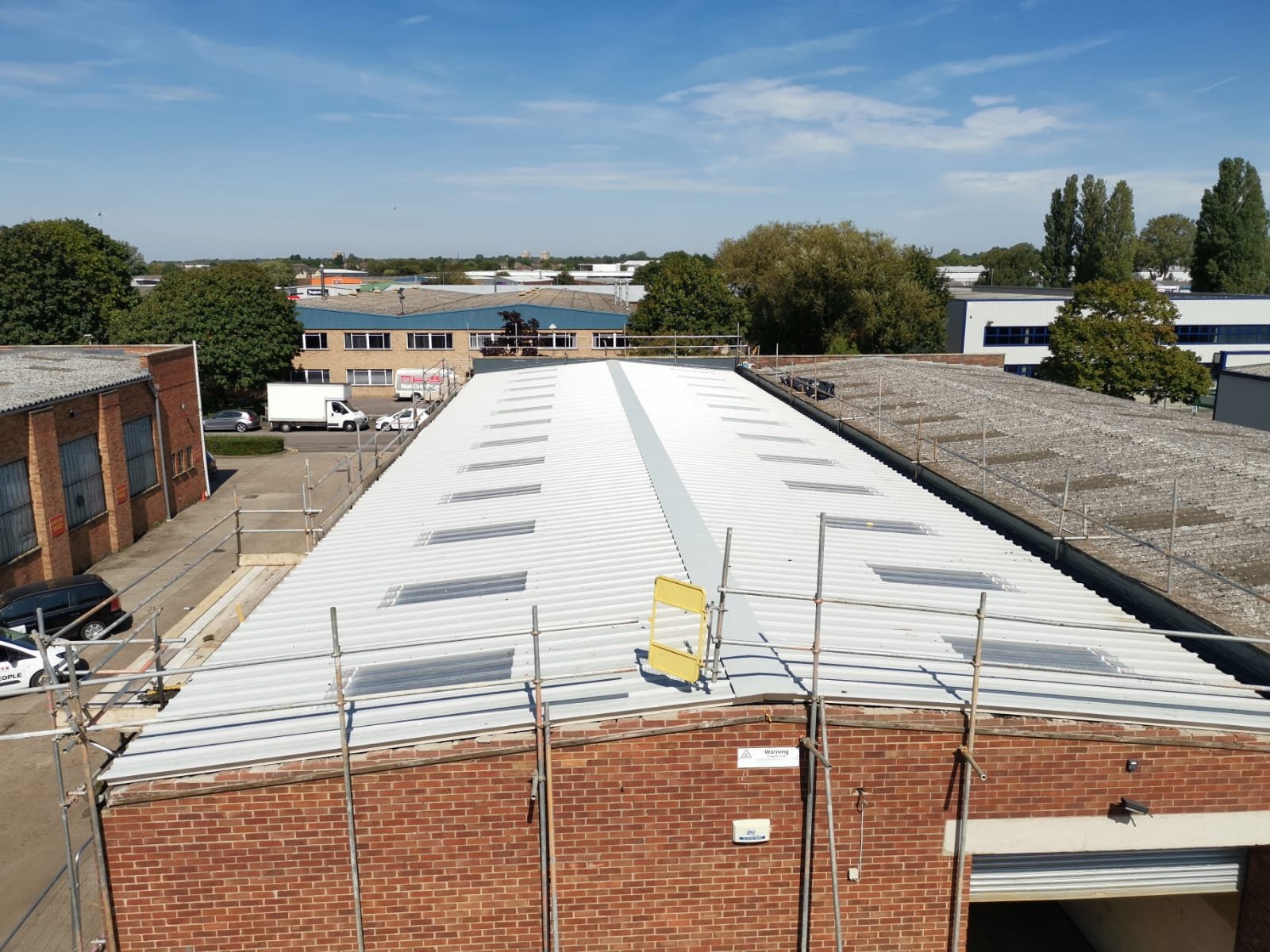 View of an industrial building rooftop with white metal roofing panels and skylights, surrounded by scaffolding, in an industrial area with trees and other buildings in the background.
