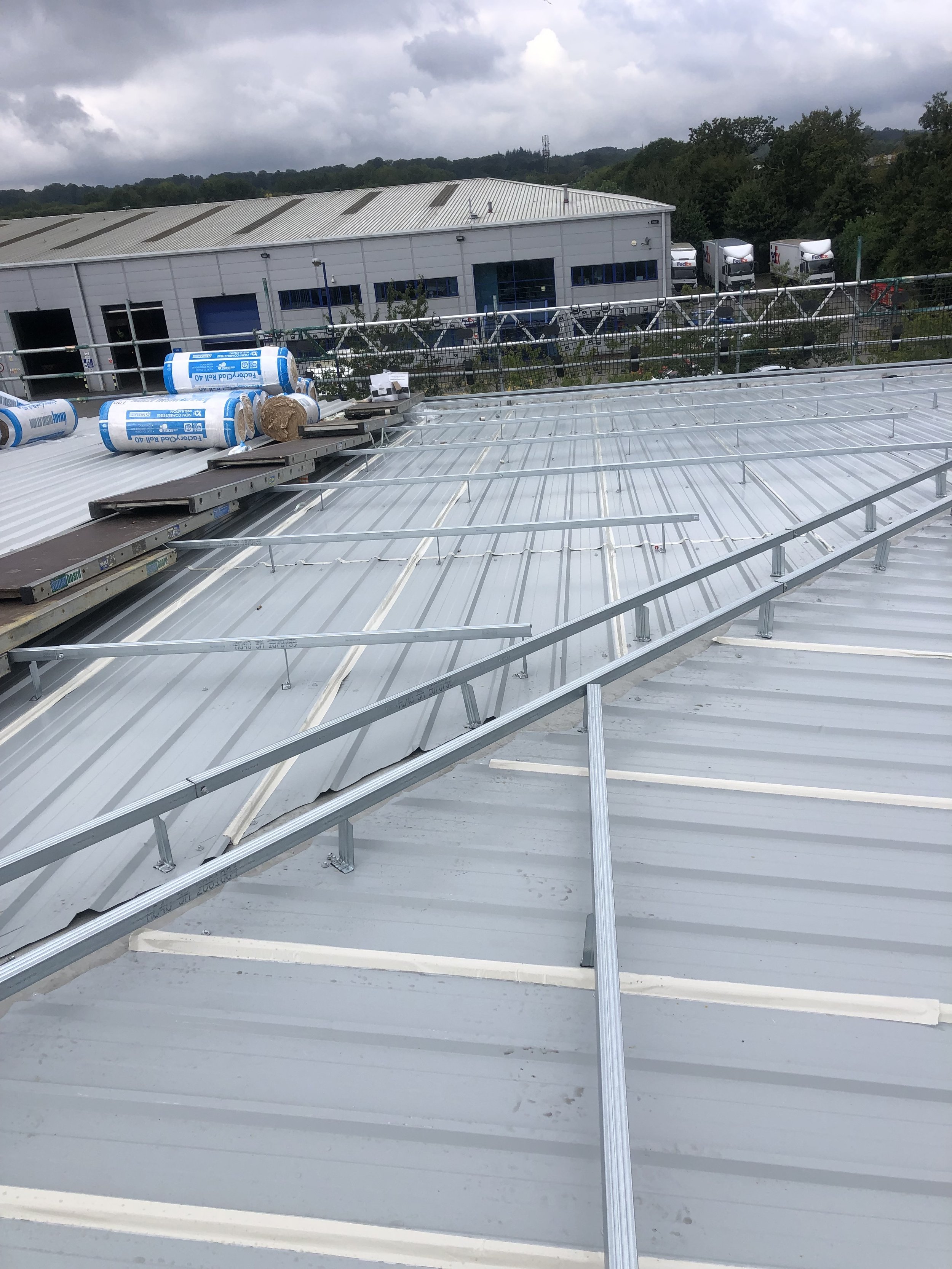 Industrial warehouse roof installation with metal sheeting and insulation rolls on rooftop; neighboring warehouse buildings and parked trucks in background.