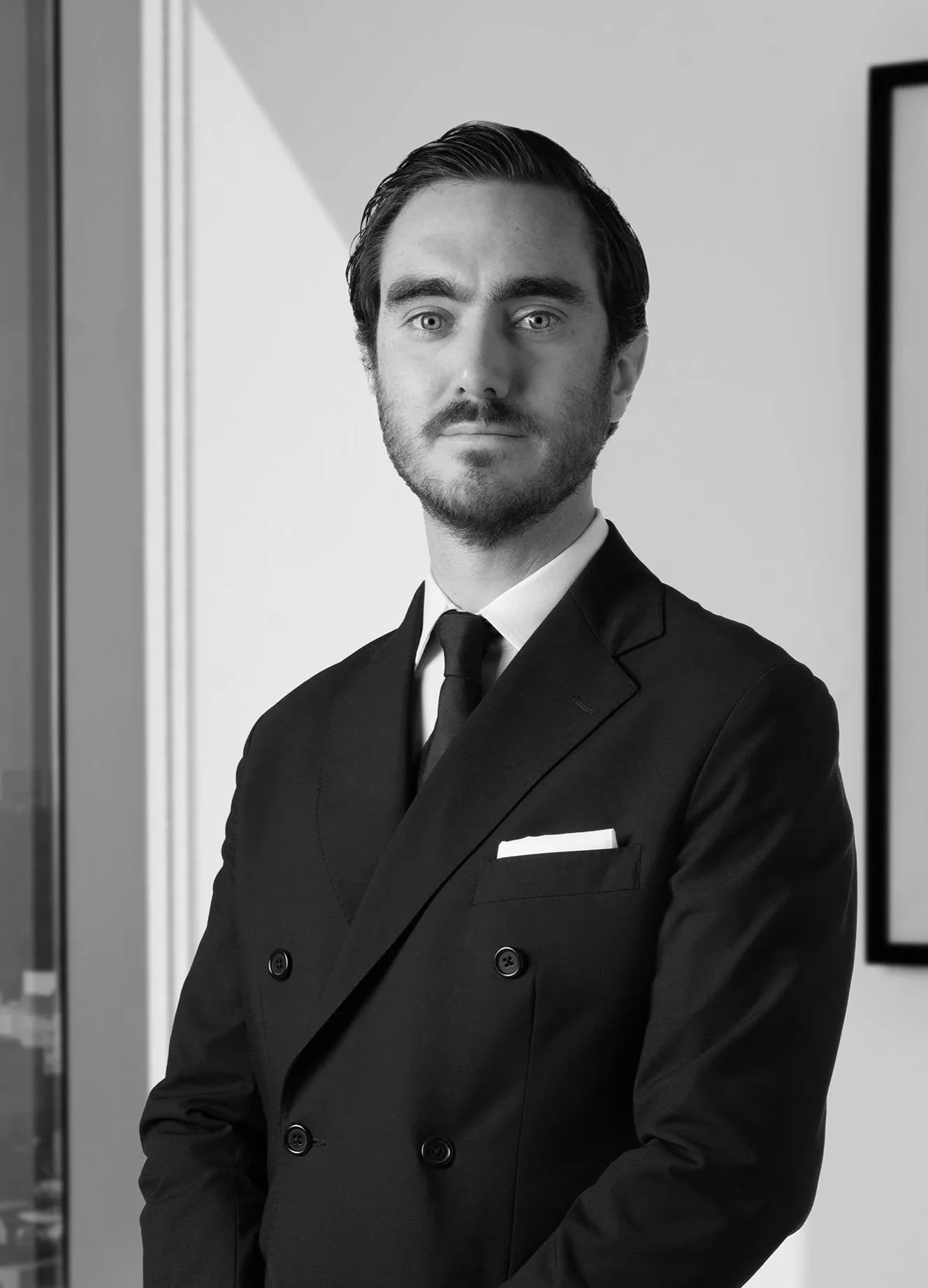 Hombre con barba y cabello peinado, vestido con traje negro y corbata, en oficina moderna.