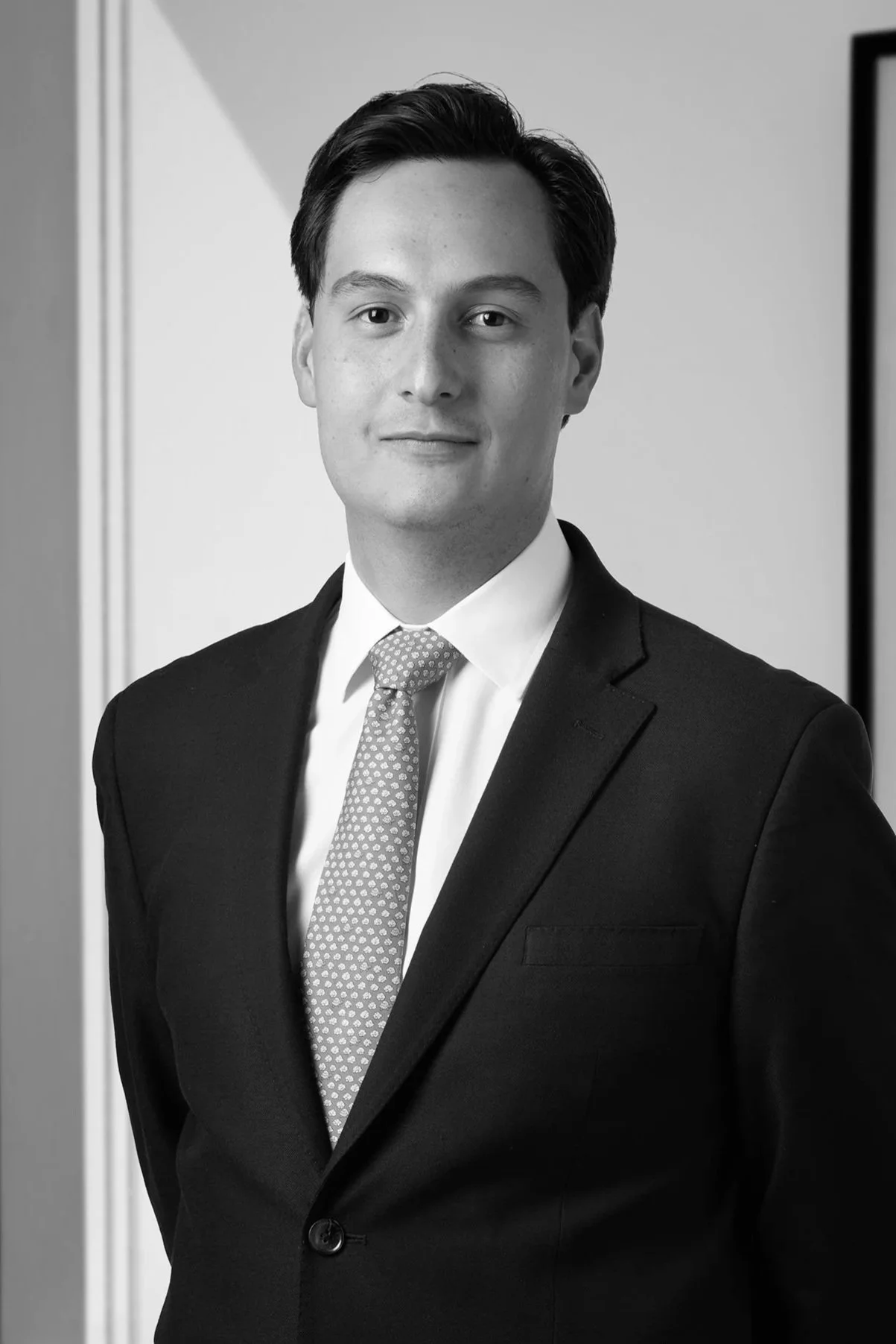 Retrato en blanco y negro de un hombre con cabello oscuro, vestido con saco negro, camisa blanca y corbata con patrón, posando de pie frente a una pared sencilla.