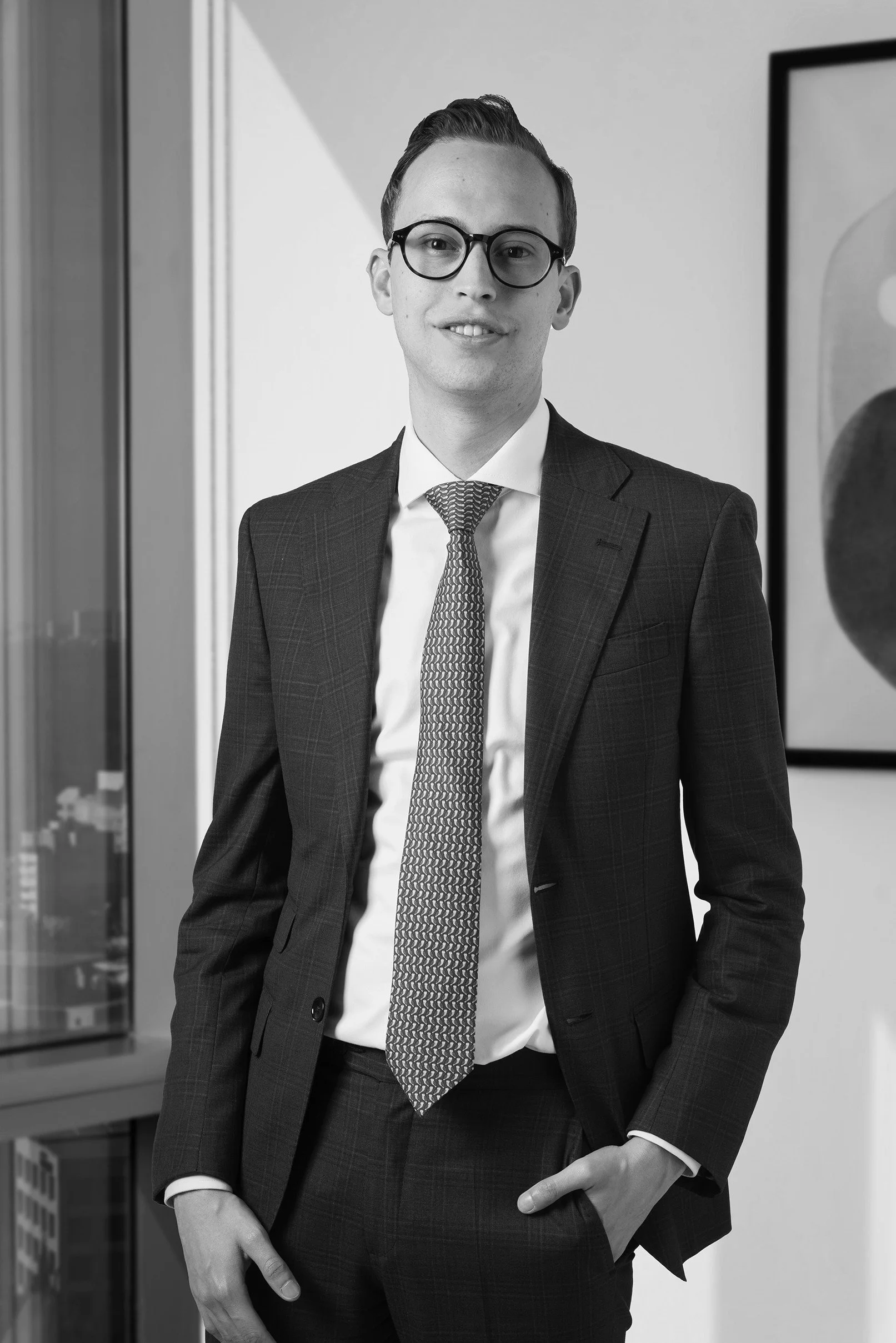 Hombre joven con gafas, vestido con traje y corbata, de pie en una oficina moderna, en blanco y negro.