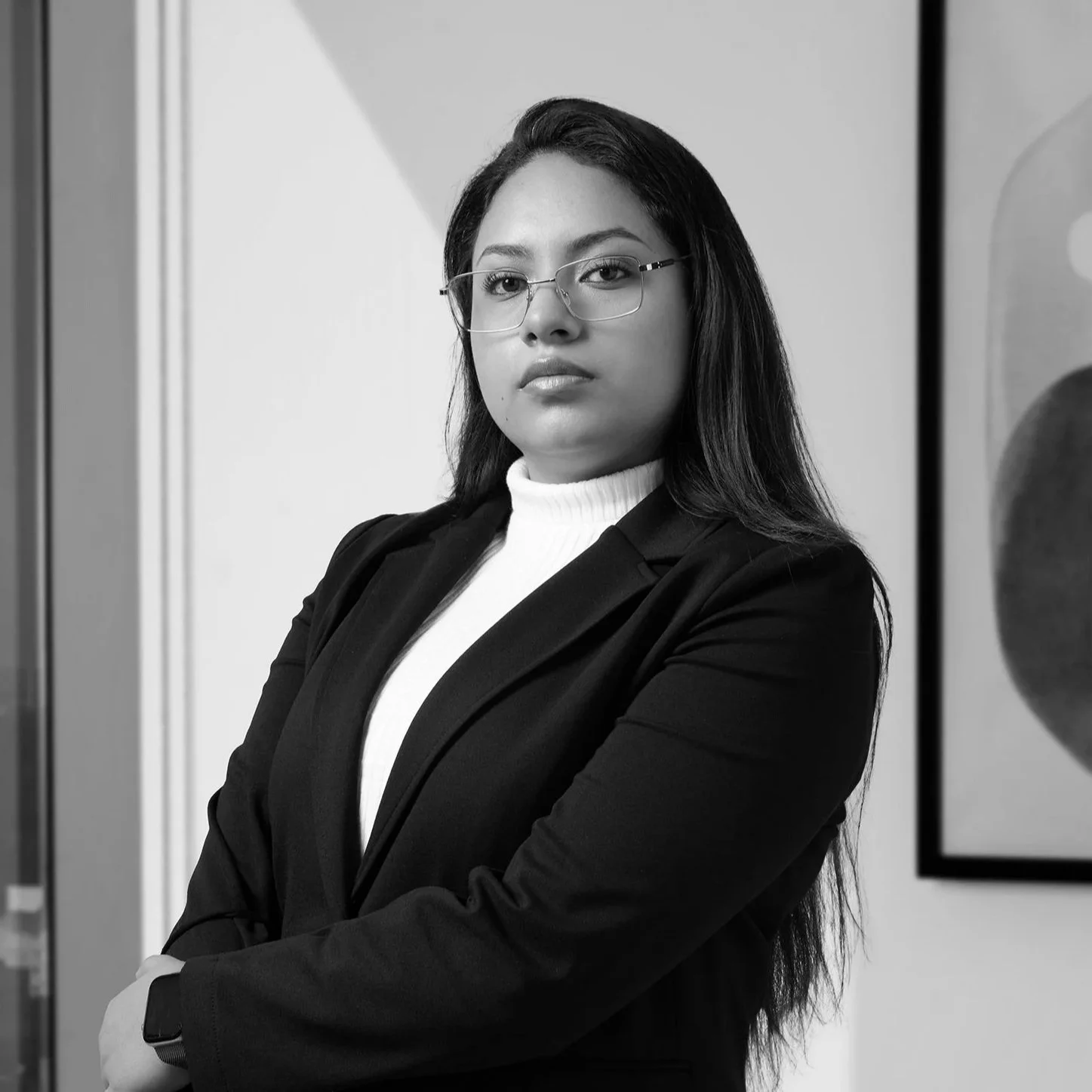 Mujer joven con gafas, vestida con blusa blanca, saco negro y pantalones blancos, posando con brazos cruzados en oficina moderna con cuadro en la pared.