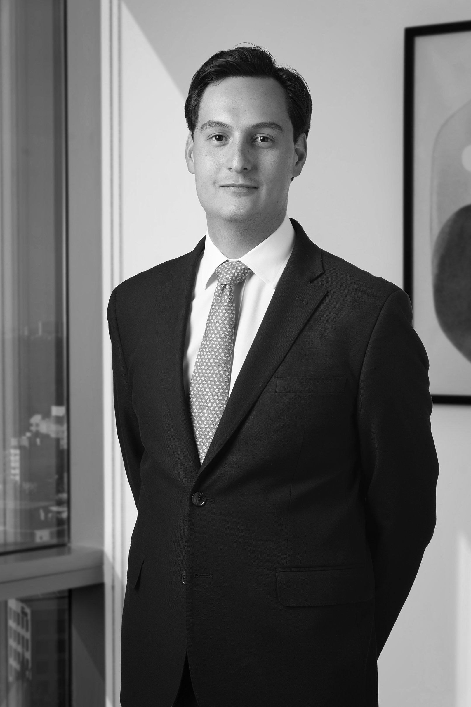 Retrato de un hombre joven con ropa formal, de cabello oscuro y peinado hacia atrás, de expresión neutral, en un entorno de oficina o espacio similar, en blanco y negro.
