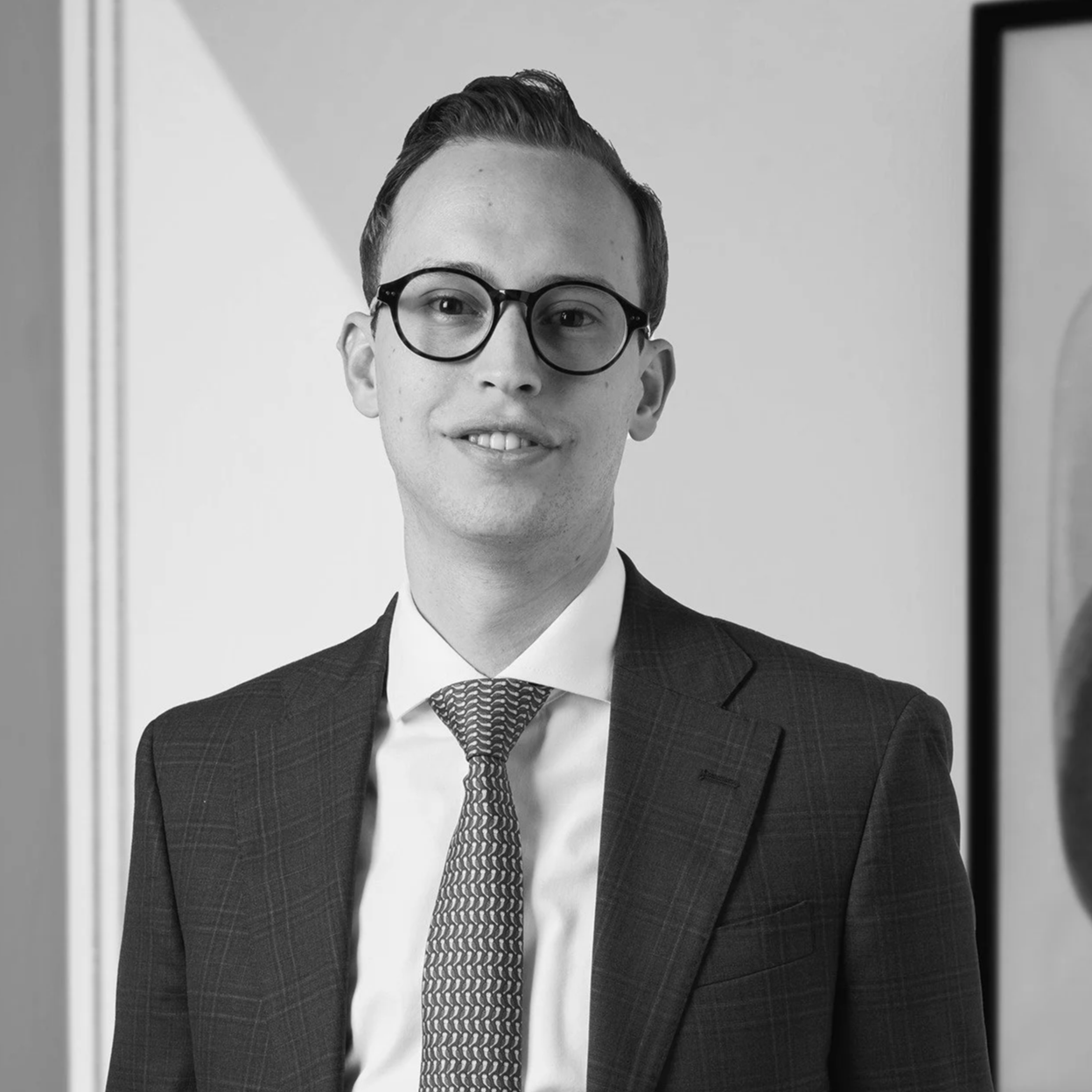 Retrato de un hombre joven con gafas, vestido con saco y corbata en un entorno profesional, en blanco y negro.