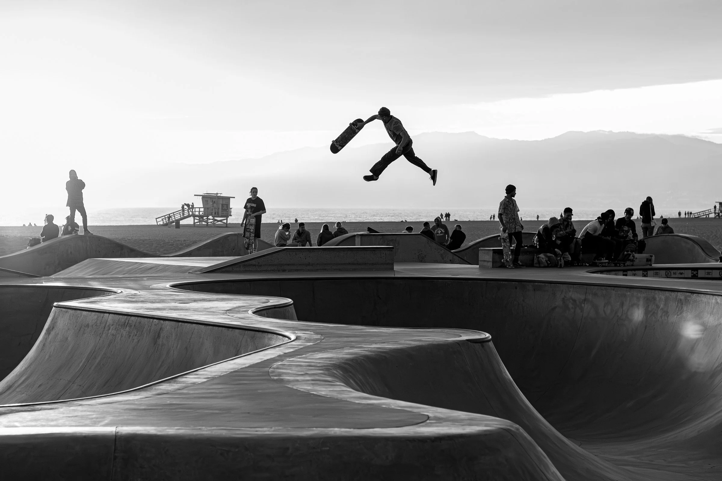 Skateboarder mid-air silhouette at Venice Beach skatepark with mountains and ocean California