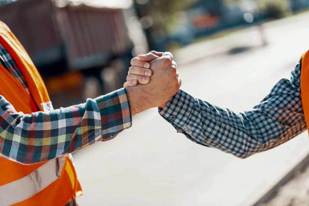 Two people in checkered shirts shake hands outdoors, both wearing safety vests, with a blurred background of a street.