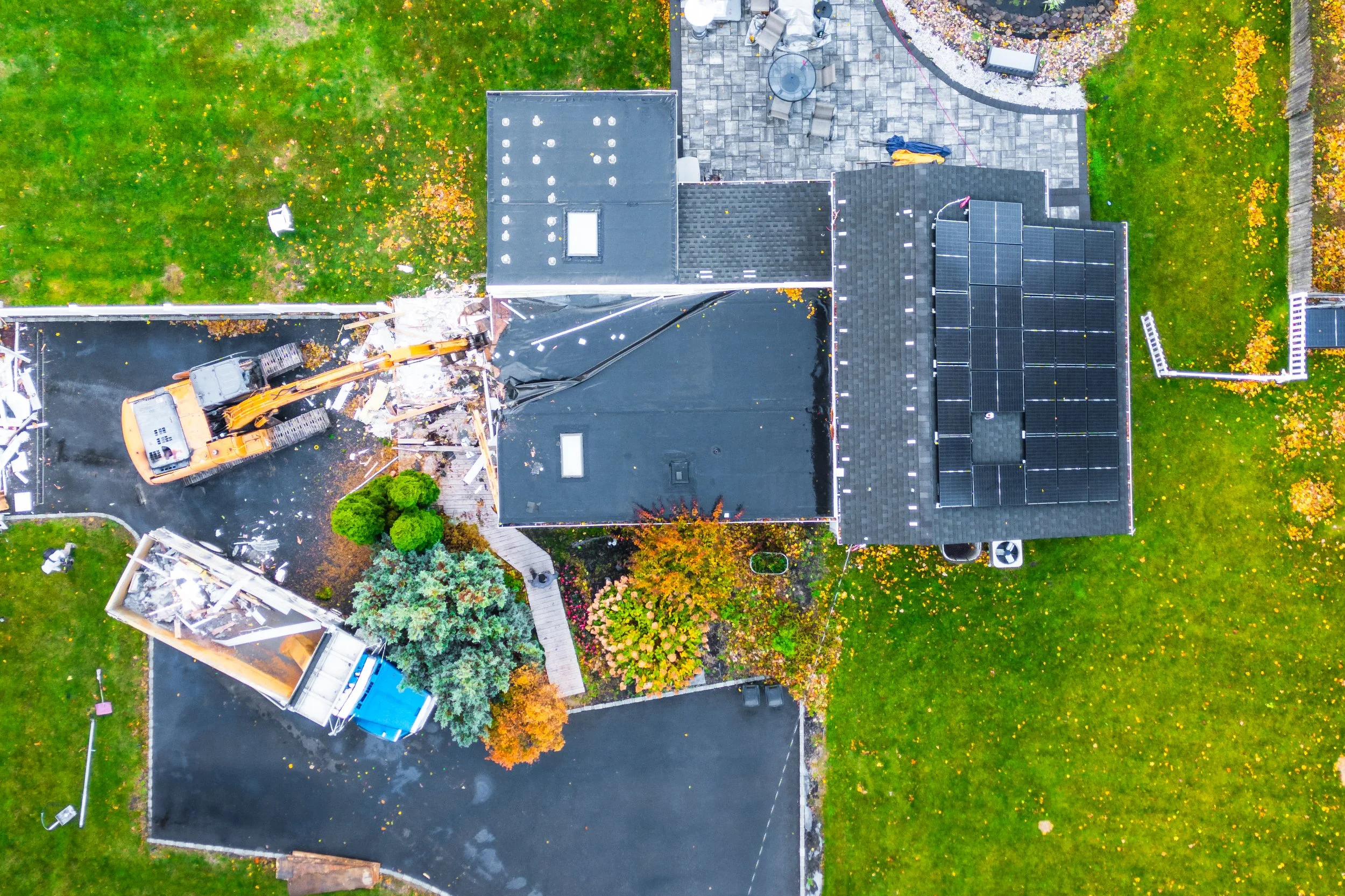 An aerial view of a house with part of the roof being broken and removed by a construction excavator. The house is surrounded by a lush green yard with scattered fall leaves, and there are construction materials and equipment nearby.