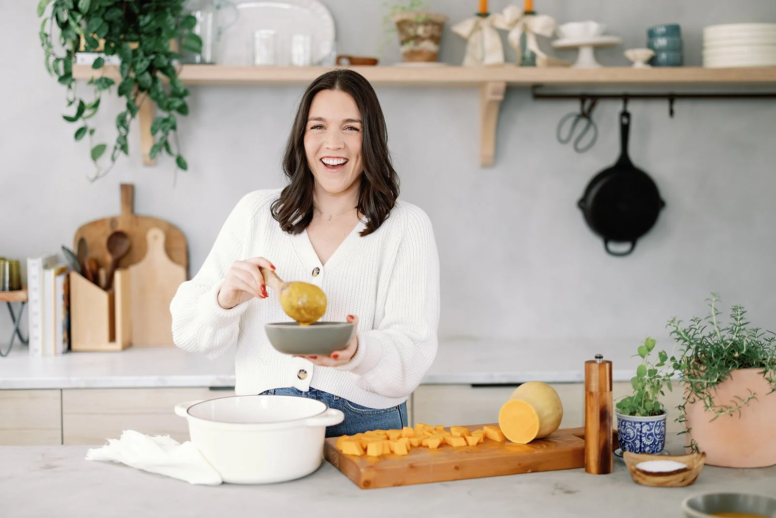 A woman in a white sweater preparing food in a modern kitchen with green plants and wooden shelves, chopping a butternut squash on a cutting board.