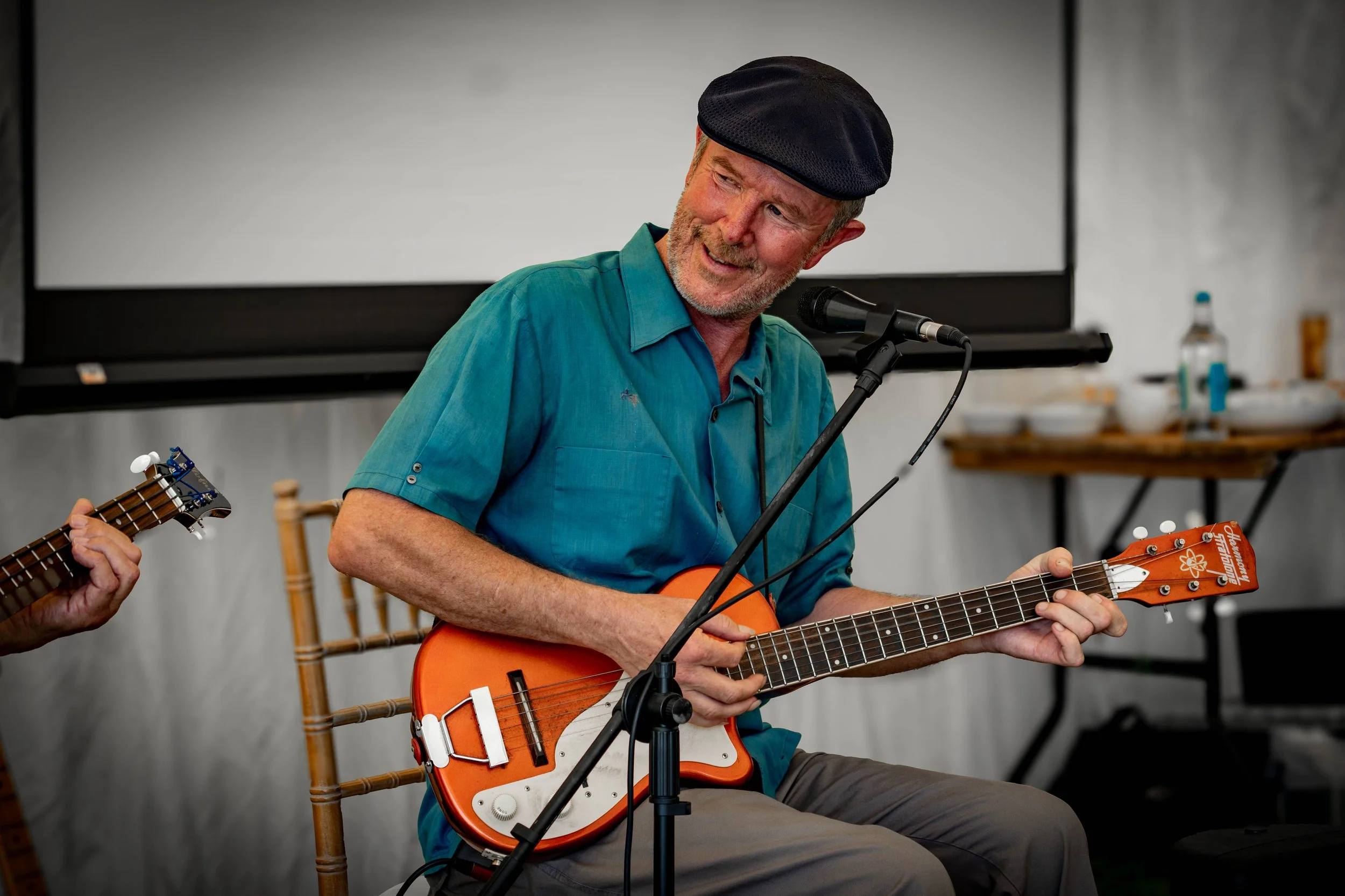 An older man wearing a black cap and teal shirt playing an orange electric ukulele while sitting on a wooden chair, with a microphone in front of him and a person playing a guitar beside him.