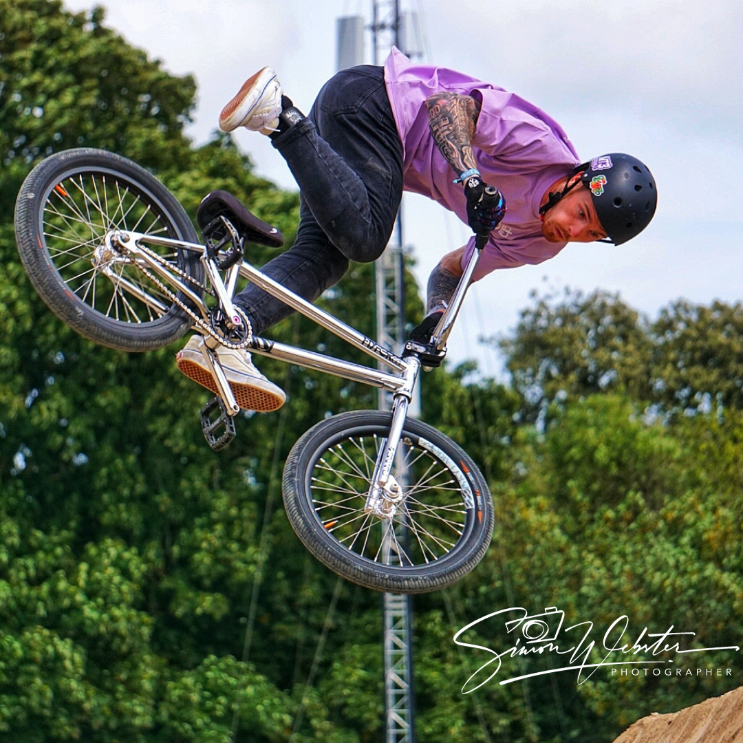 A man performing BMX trick in mid-air, wearing a black helmet, purple shirt, black jeans, and sneakers, with tattoos on his arms and a green outdoor background.