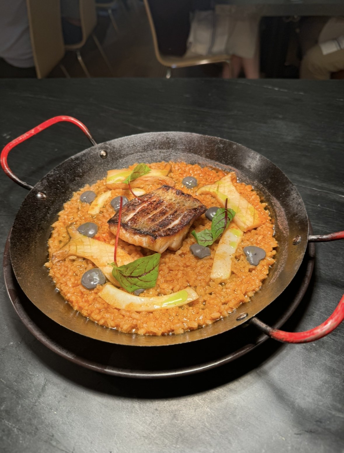 A pan of risotto garnished with grilled fish, green leaves, and small gray beans.