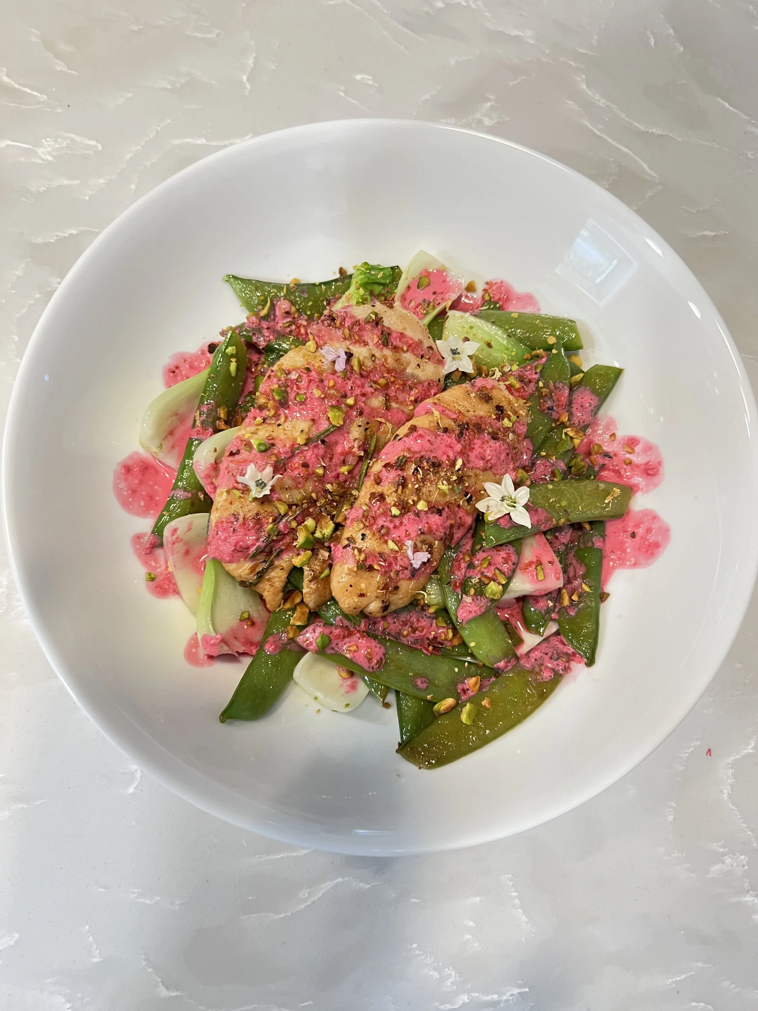 Fresh salad with green beans, sliced vegetables, topped with grilled chicken, pink sauce, chopped nuts, and edible flowers in a white bowl.