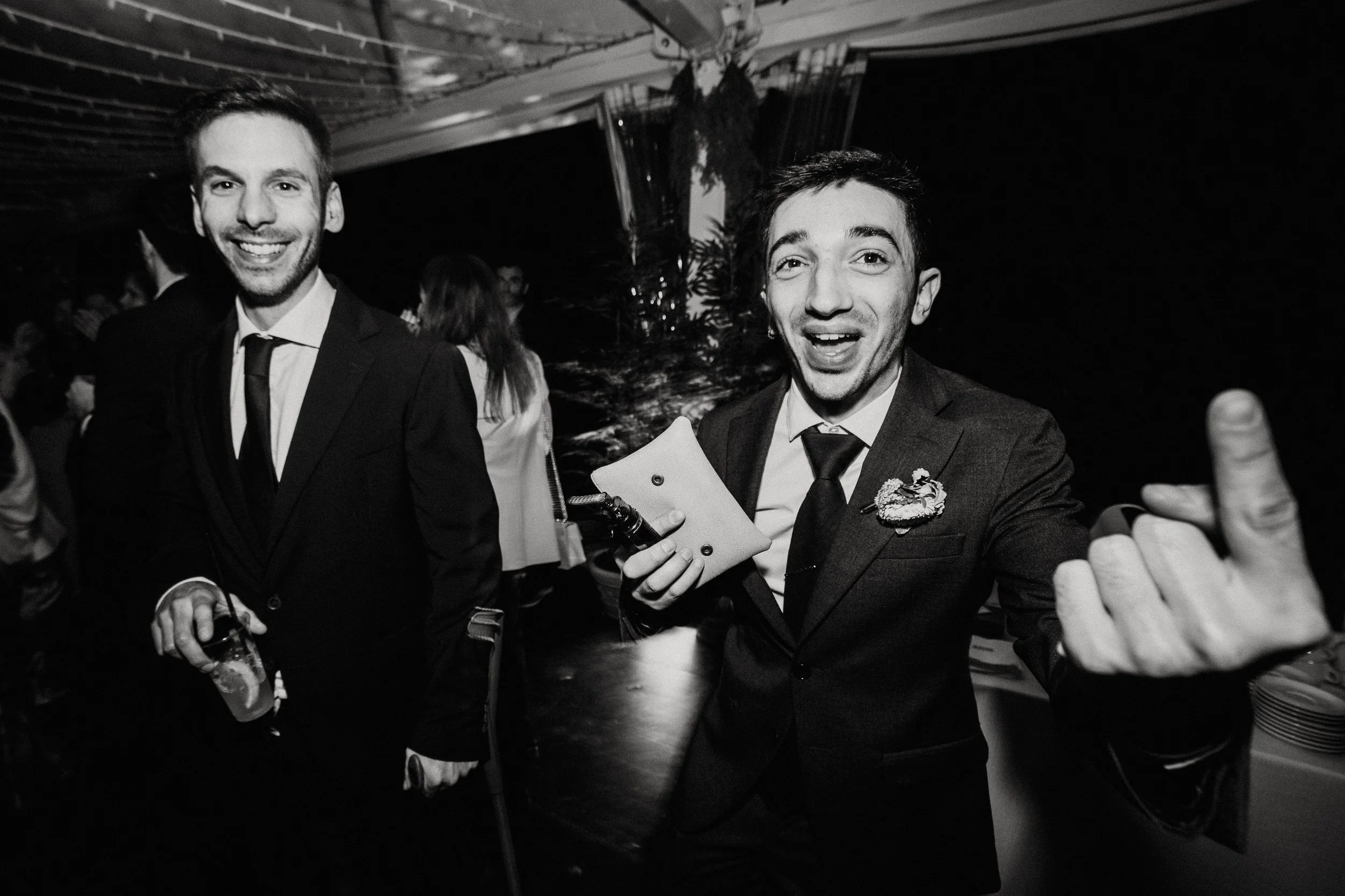 Due uomini sorridenti in abiti eleganti durante un evento sociale o festa, con uno che fa un gesto con la mano.