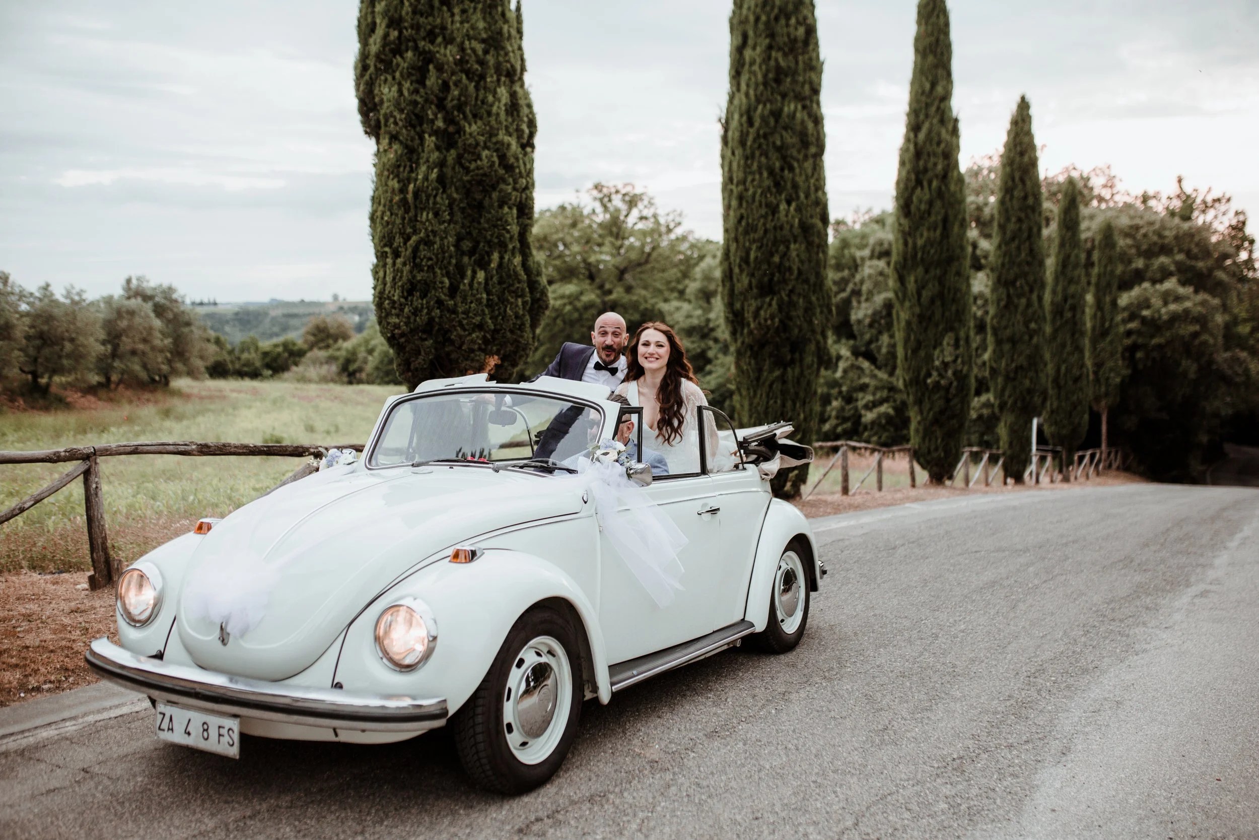 Una coppia felice in abiti da sposa e sposo in un'auto decappottabile bianca, sulla strada rurale tra cipressi.