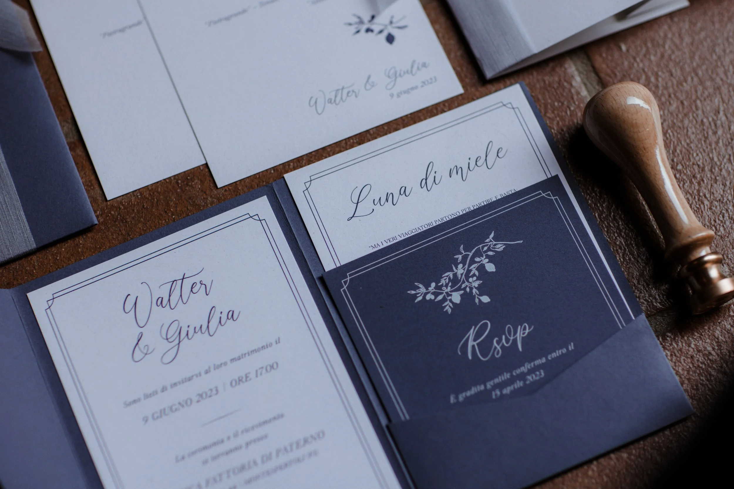 Biglietti di invito e menu per un matrimonio, con tema blu e decorazioni floreali, e uno spillino di legno accanto.