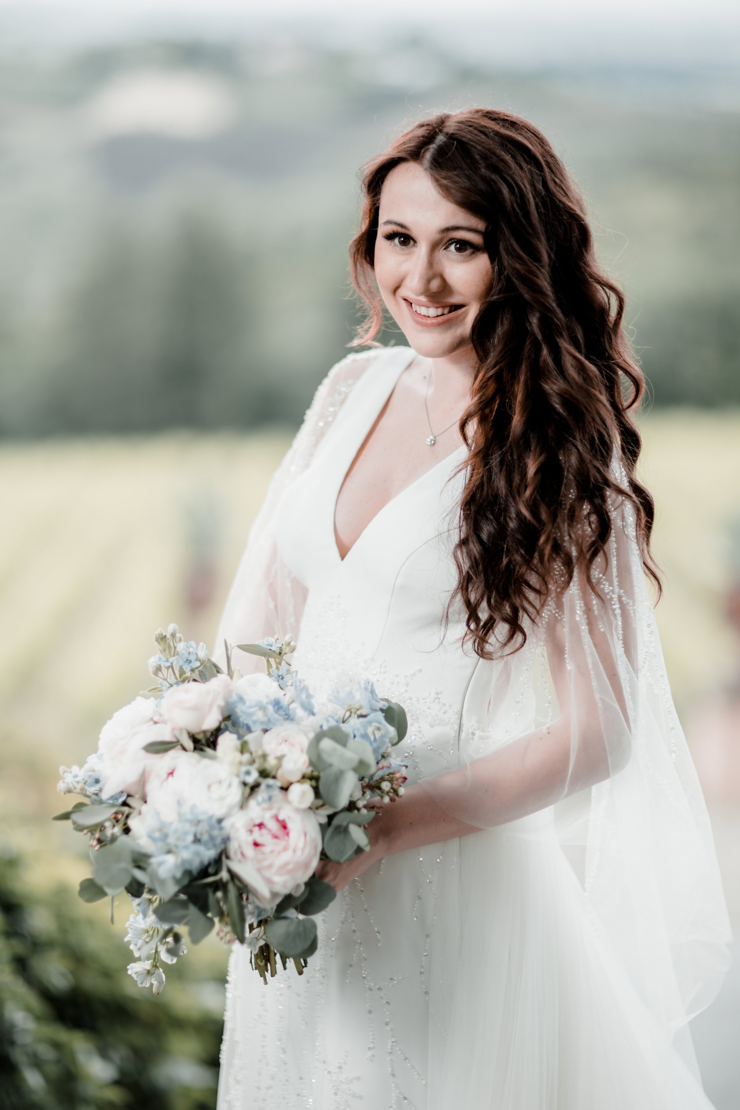 Una donna in abito da sposa con boccoli castani scuri, che sorride e tiene un mazzo di fiori pastello, in un paesaggio naturale con sfondo sfocato.