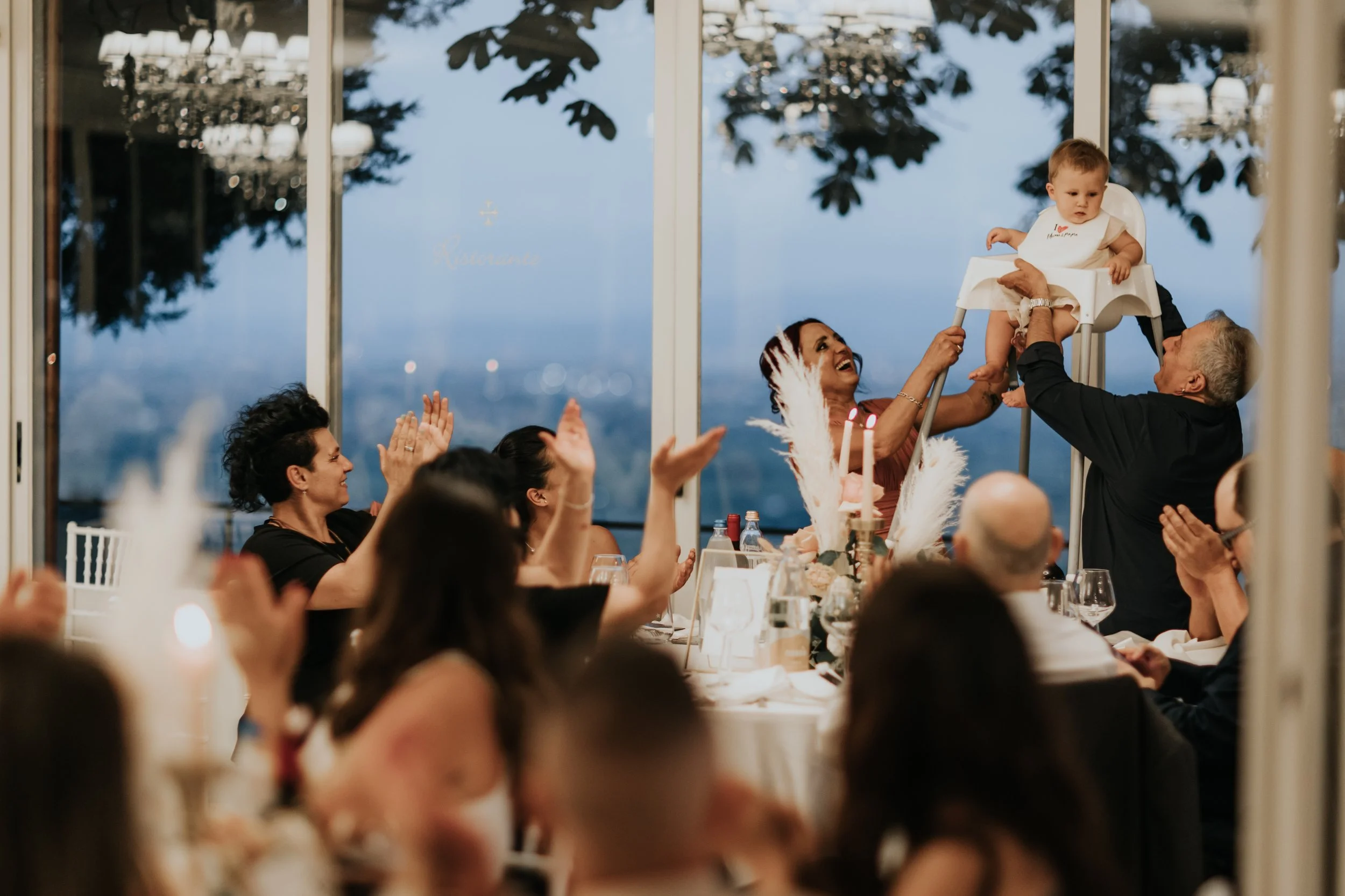 Una famiglia festeggia un compleanno con un bambino seduto in una sedia alta, circondato da adulti che applaudono e sorridono in un ristorante con viste sulla città al tramonto.