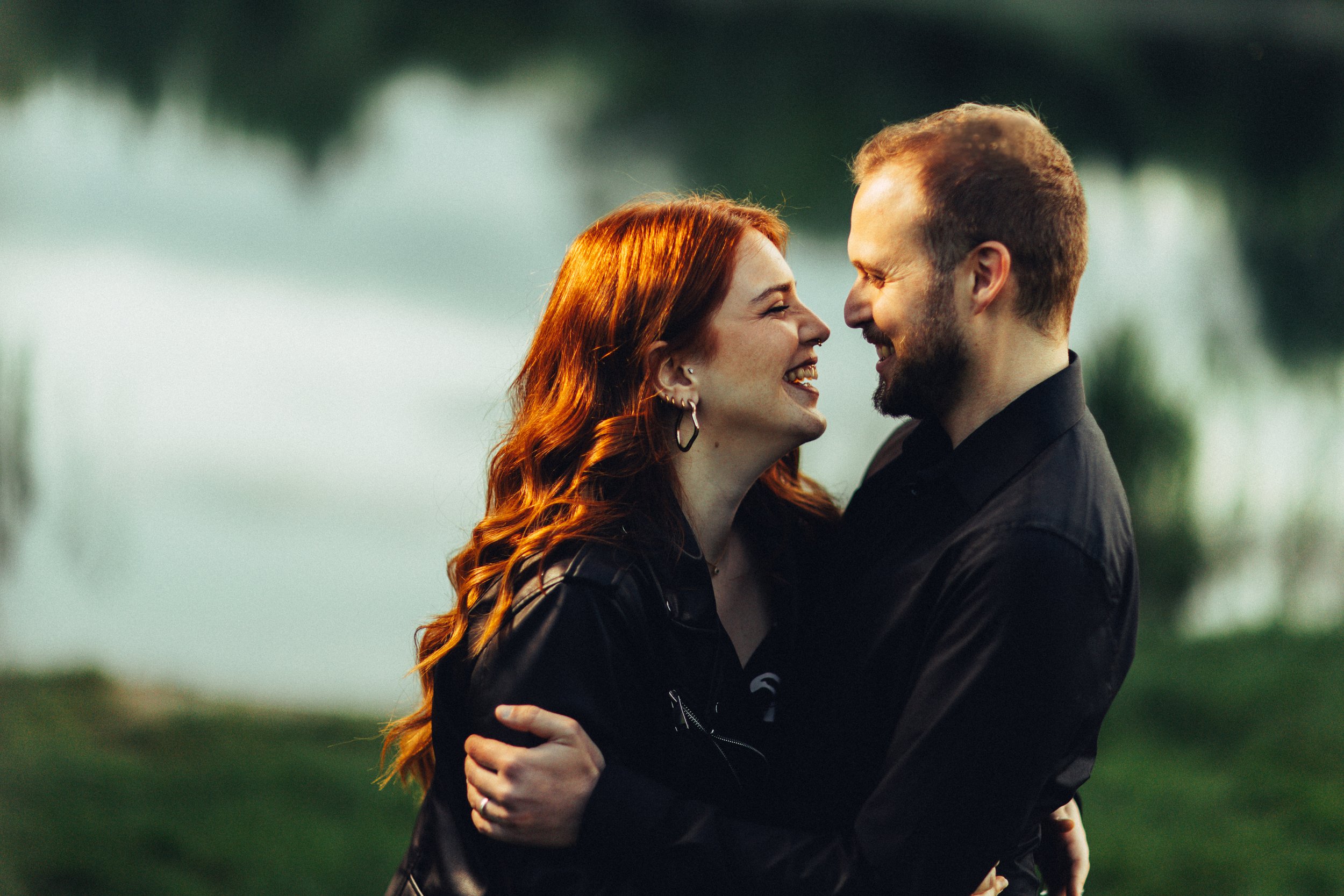 Una coppia felice che sorride e si abbraccia all'aperto vicino a un corpo d'acqua, con uno sfondo naturale e alberi.